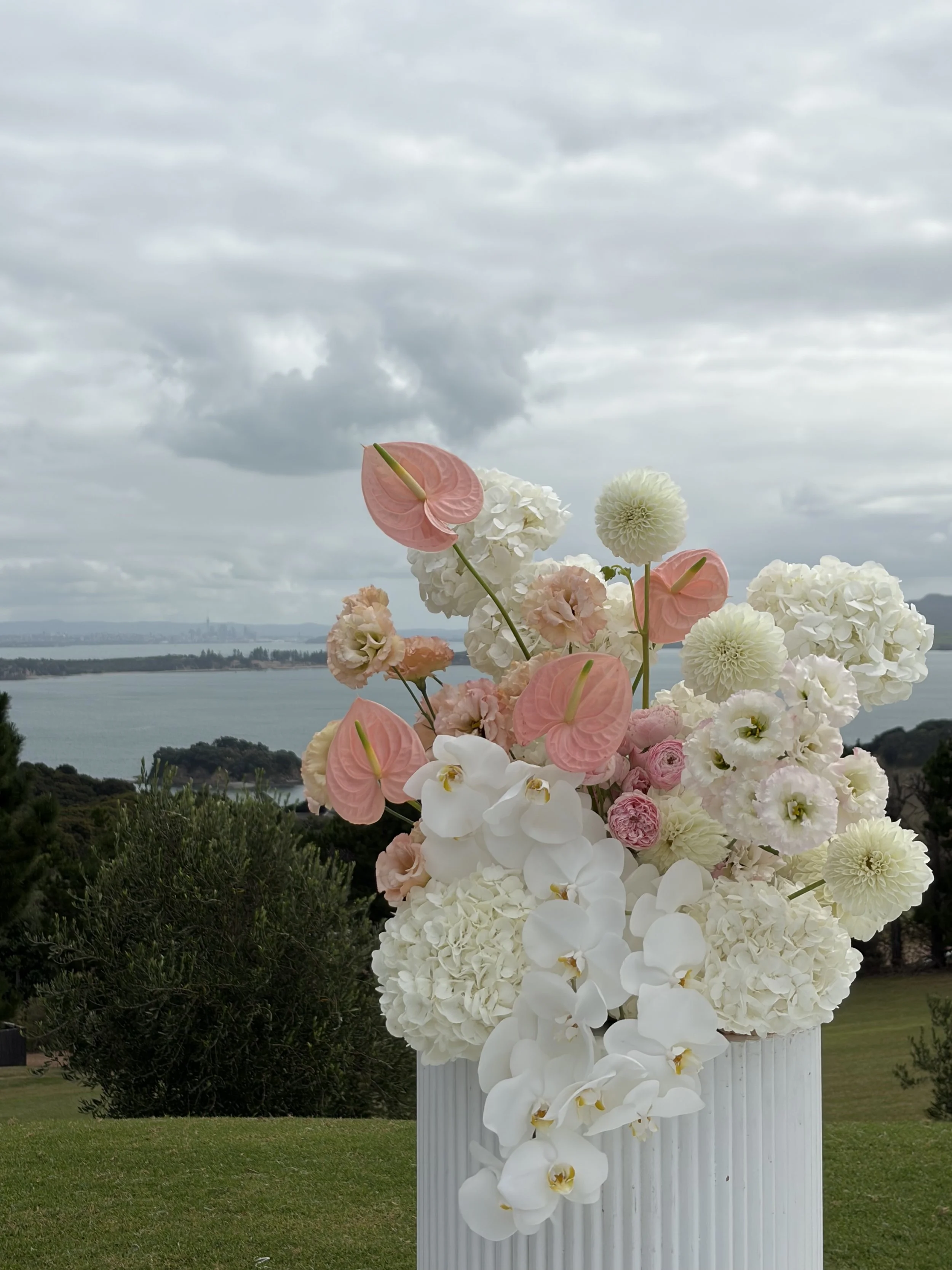 Vase of pink and white flowers including orchids and hydrangeas outdoors with a cloudy sky and water in the background.
