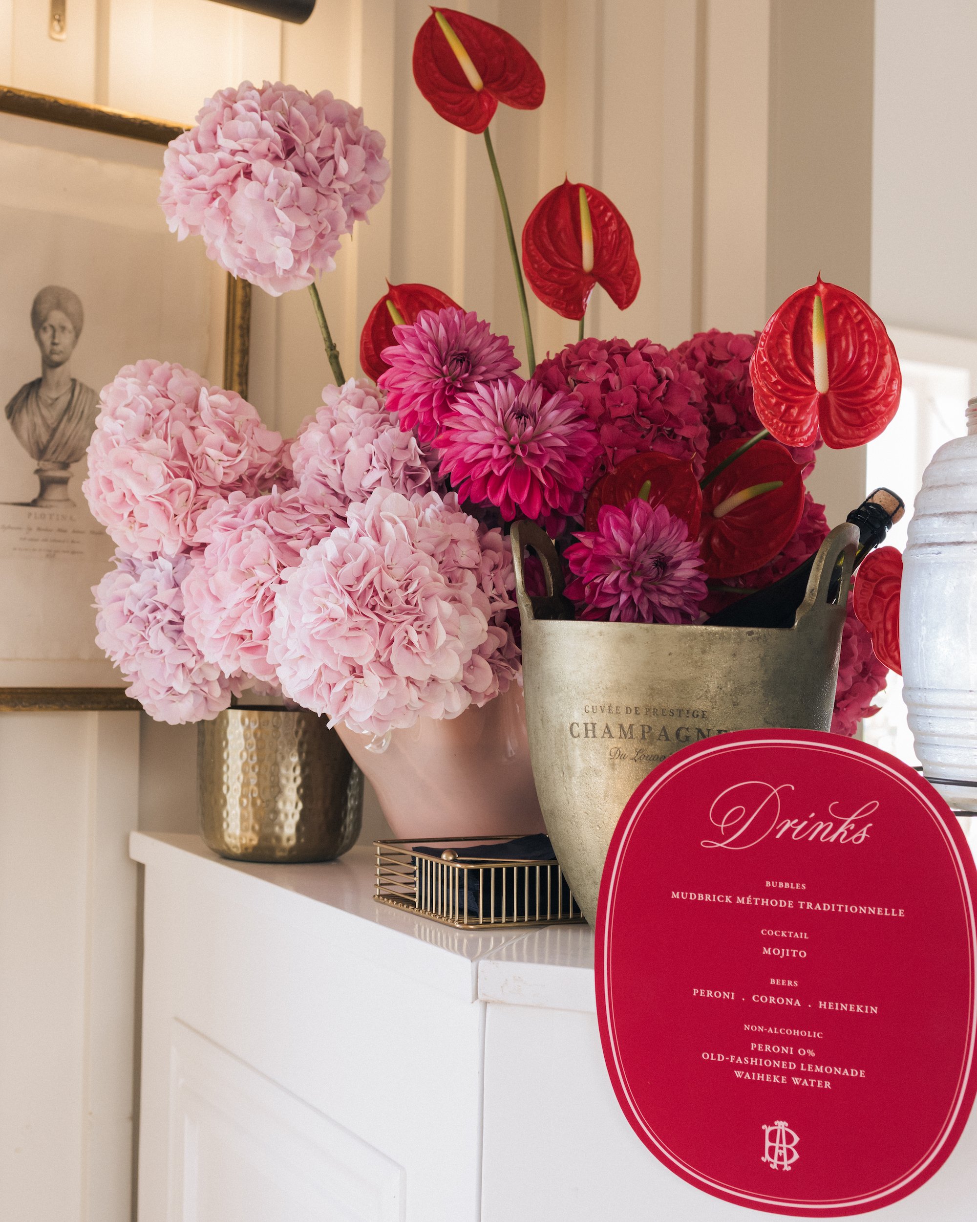 A bouquet of pink hydrangeas, pink dahlias, and red anthuriums placed in a decorative black and gold vase on a white shelf. In the background, a vintage-style framed black-and-white portrait of a woman wearing a dress and hat. To the right, part of a
