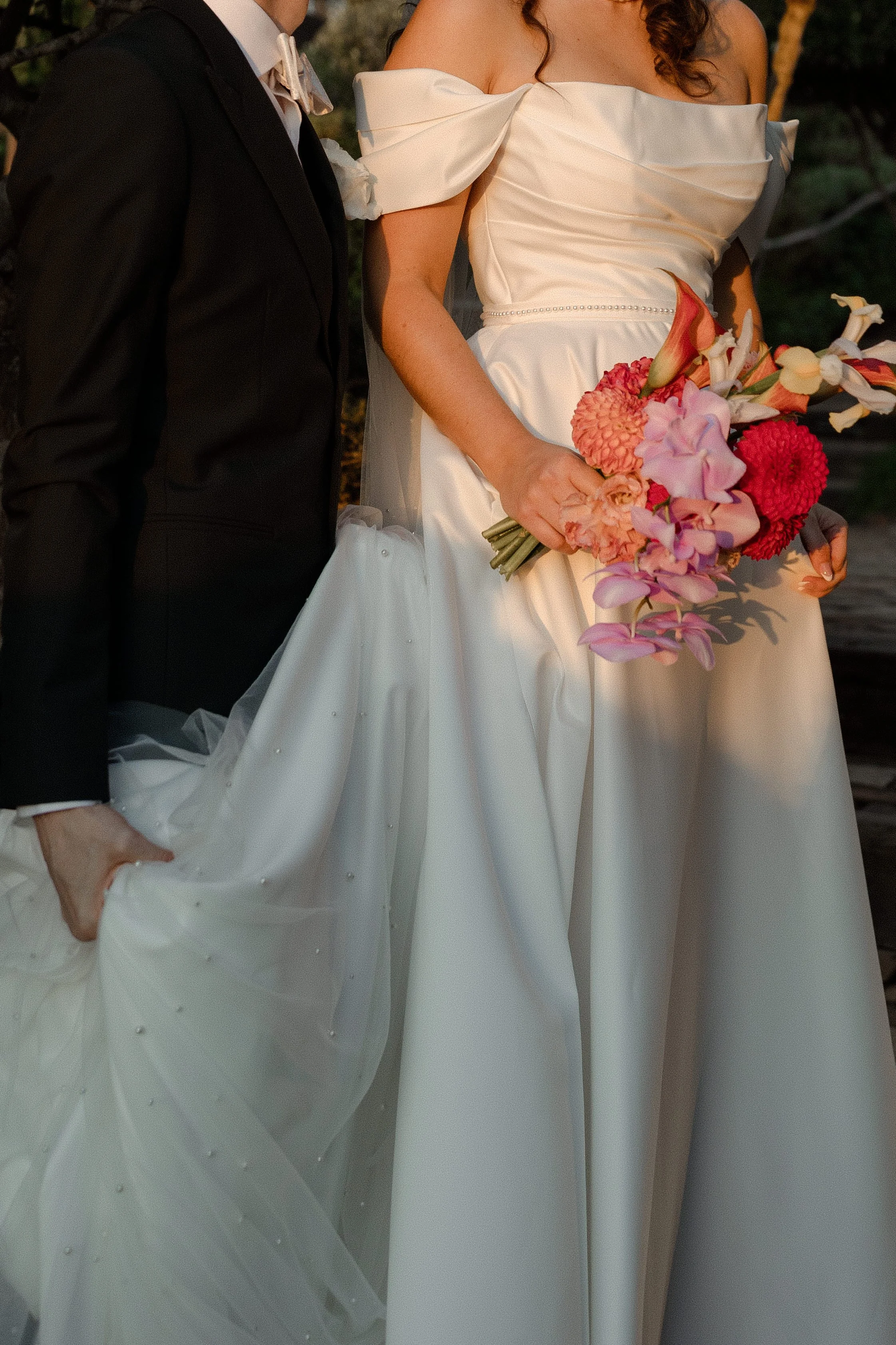 Close-up of a bride in a white wedding gown holding a colorful bouquet of flowers, standing beside a groom dressed in a black suit and bowtie, during a wedding ceremony.