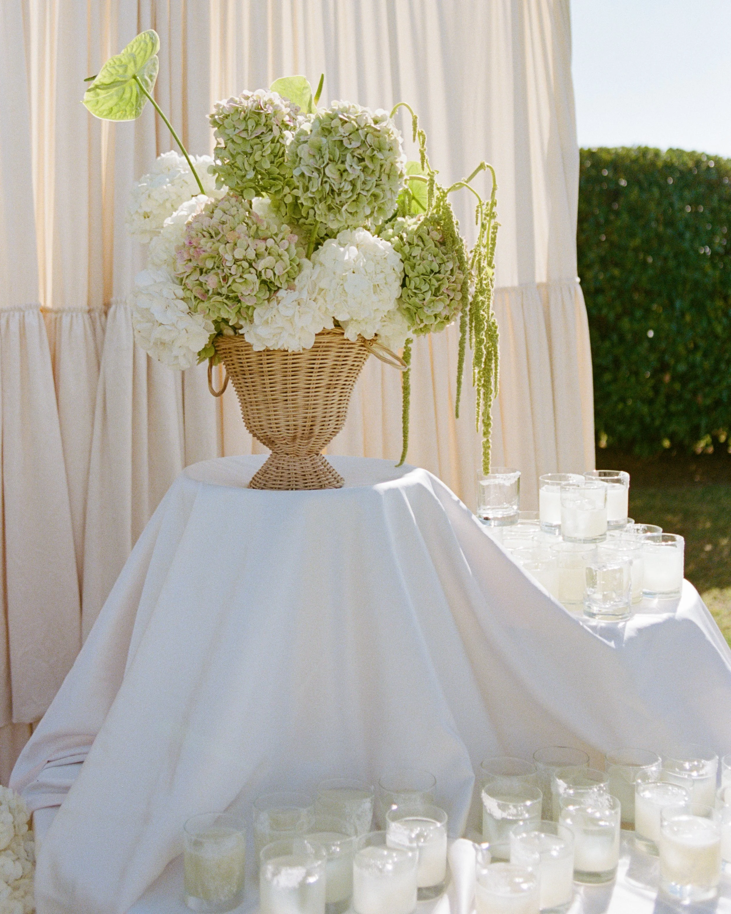 A floral arrangement with white and light green hydrangeas and an anthurium in a wicker basket on a white table, set against a neutral backdrop with cream curtains and outdoor greenery.