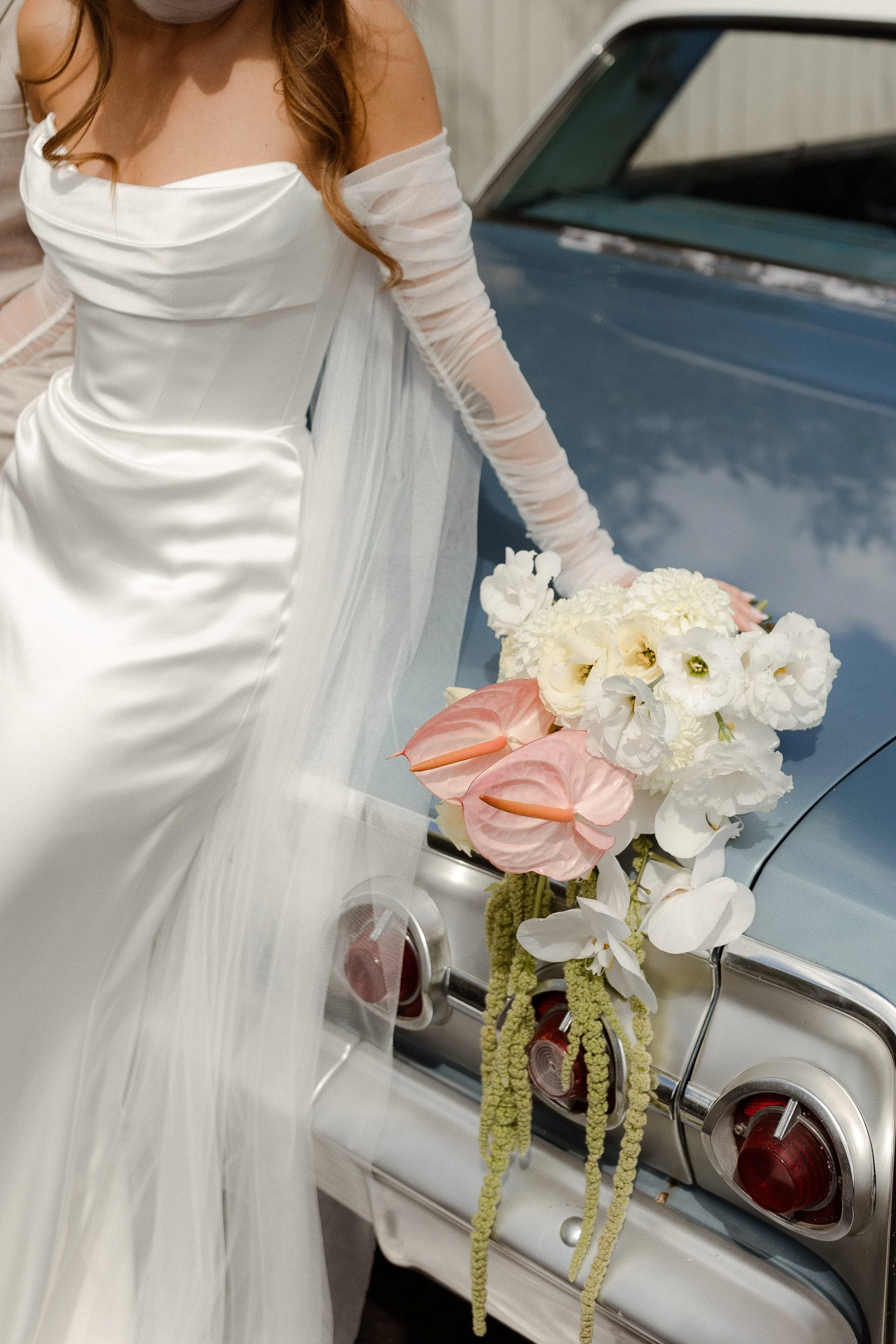 A bride in a white wedding gown and long sleeve dress stands next to a vintage car decorated with white and pink flowers, with her hand resting on the car.