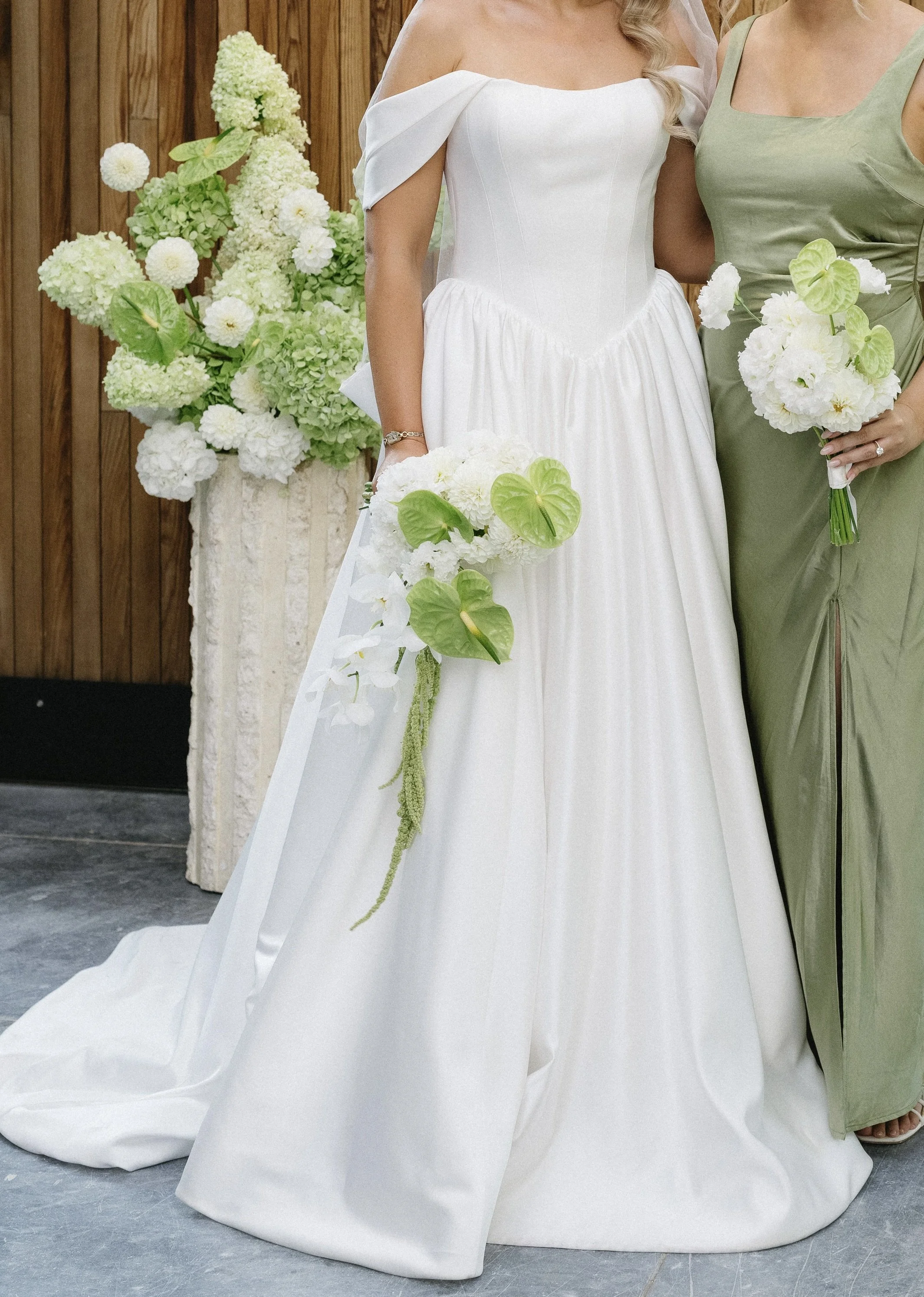 Close-up of two women in wedding dresses holding bouquets of white flowers, standing in front of a wooden wall with a large floral arrangement.