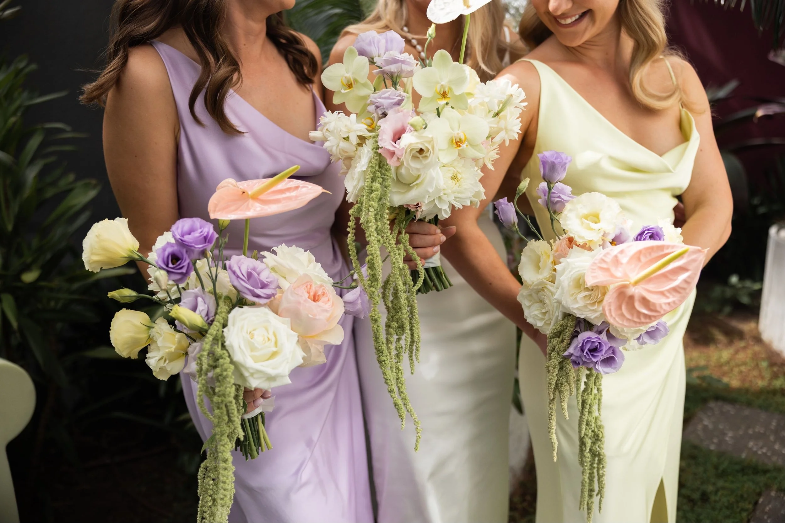 Three women in pastel-colored dresses holding bouquets of flowers, smiling at an outdoor event.