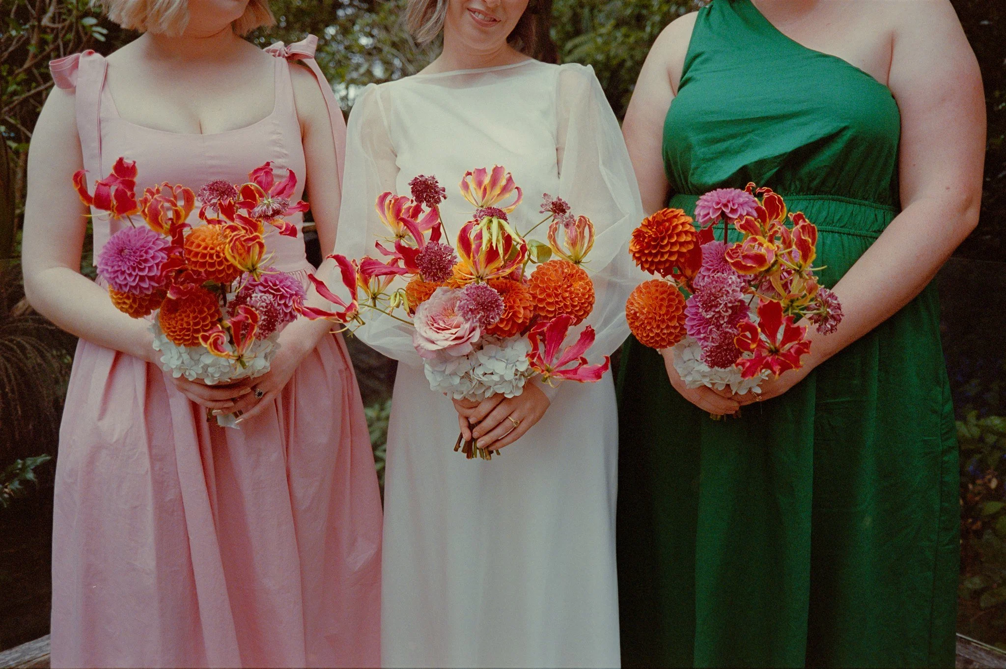 Three women in colorful dresses holding bouquets of mixed pink, orange, and red flowers outdoors.