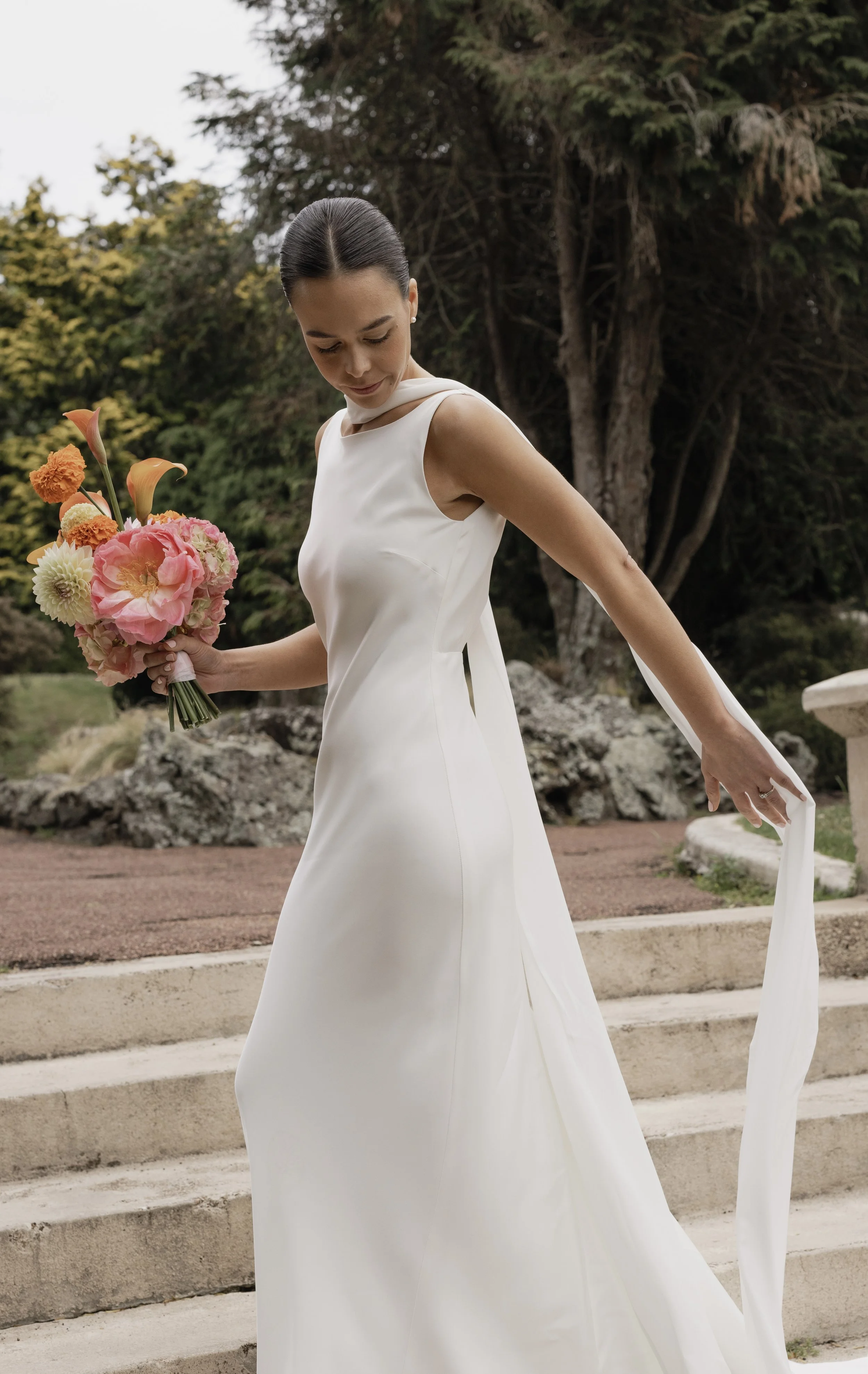 A woman in a white wedding dress holding a bouquet of flowers, walking outdoors with trees and steps in the background.