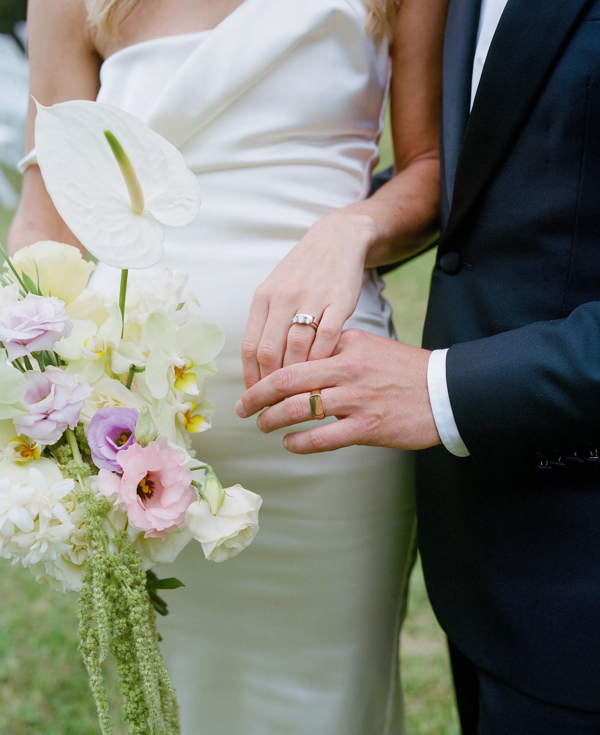Close-up of a bride and groom holding hands during a wedding, with the bride wearing a white dress, a ring on her finger, and a bouquet of white and pink flowers. The groom is wearing a dark suit and white shirt.