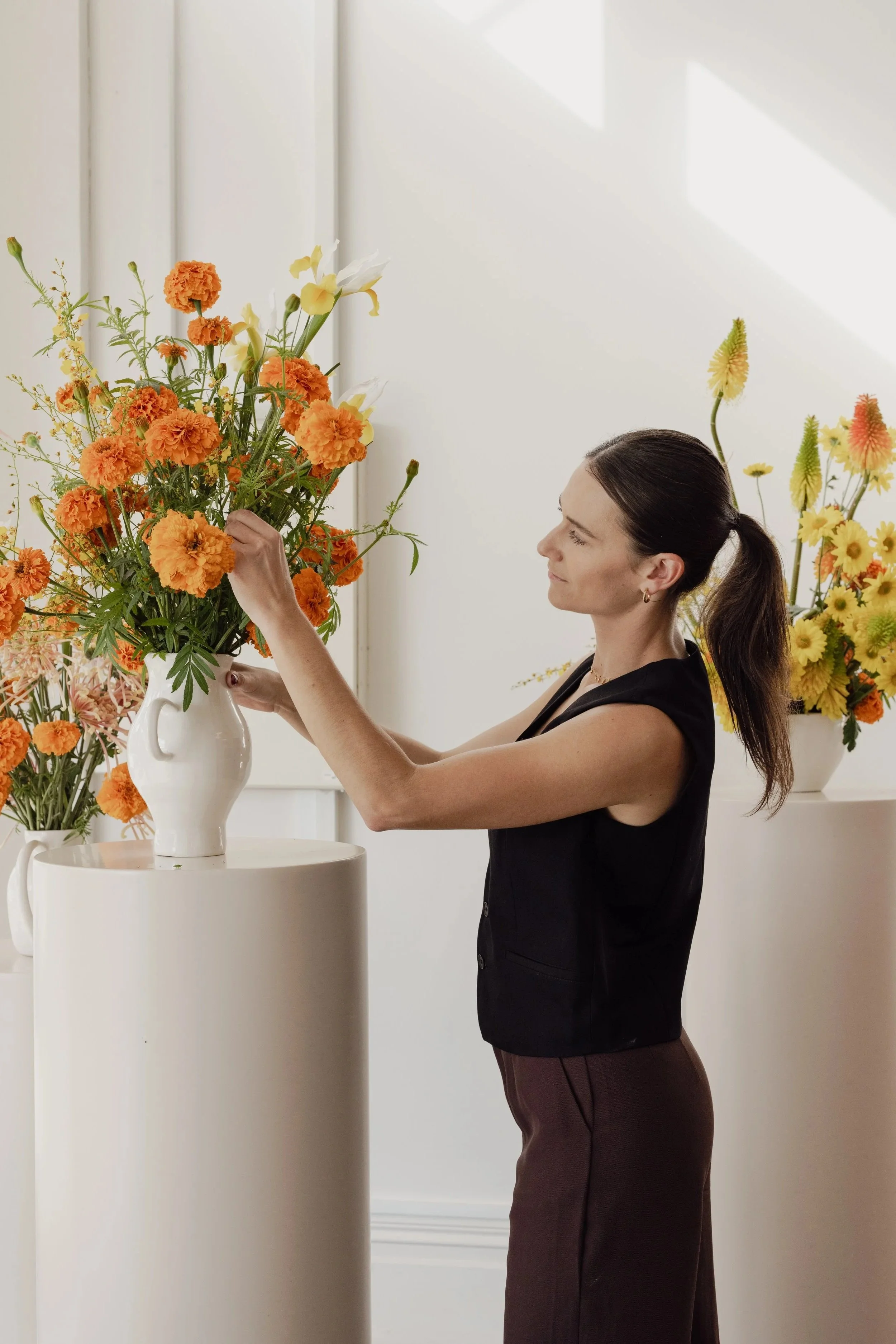 A woman with dark hair tied in a ponytail, wearing a sleeveless black top and brown pants, arranging orange and white flowers in a white vase on a round white pedestal.