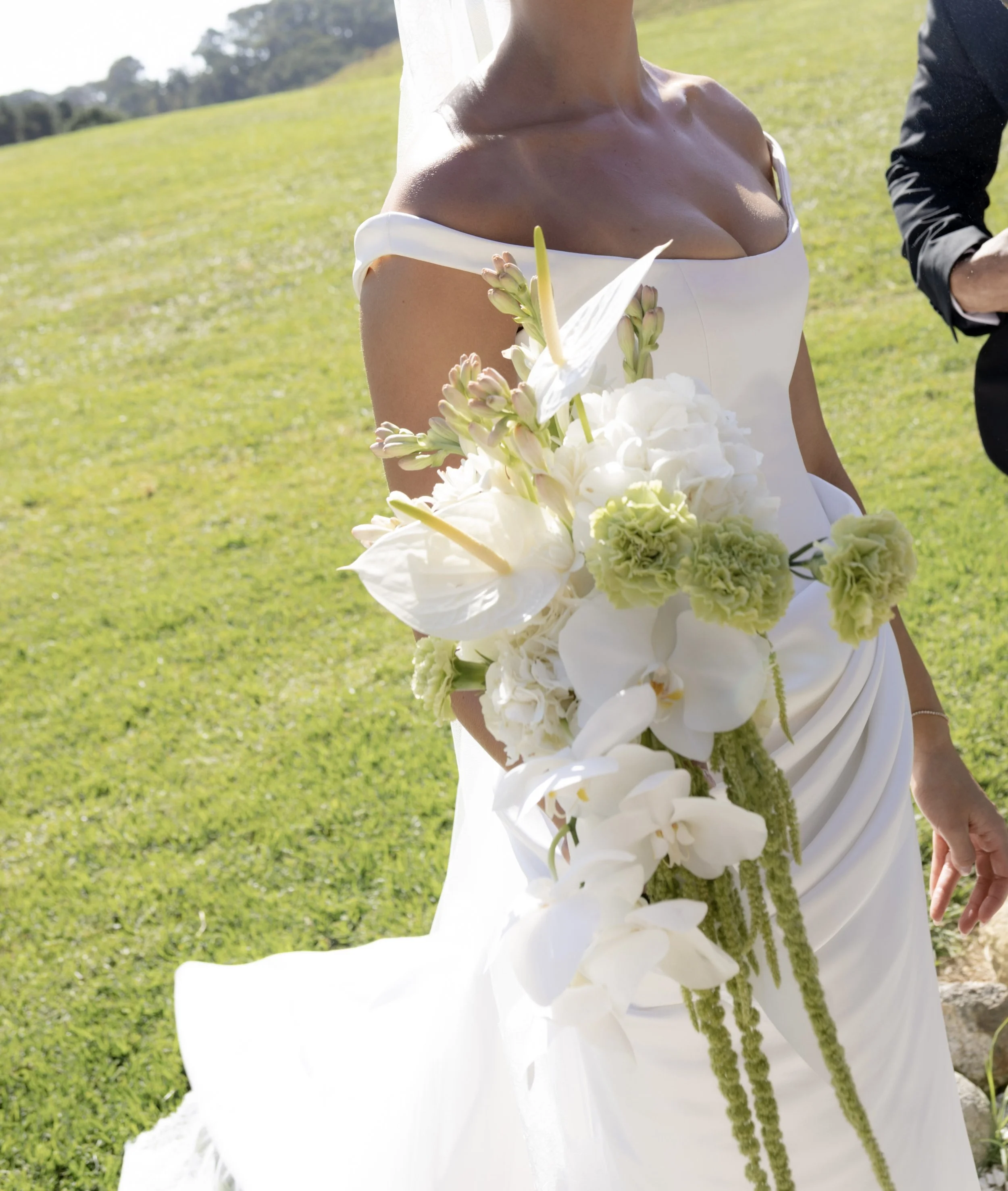 Close-up of a bride wearing an off-the-shoulder white wedding gown holding a bouquet of white flowers outdoors with a grassy field in the background.