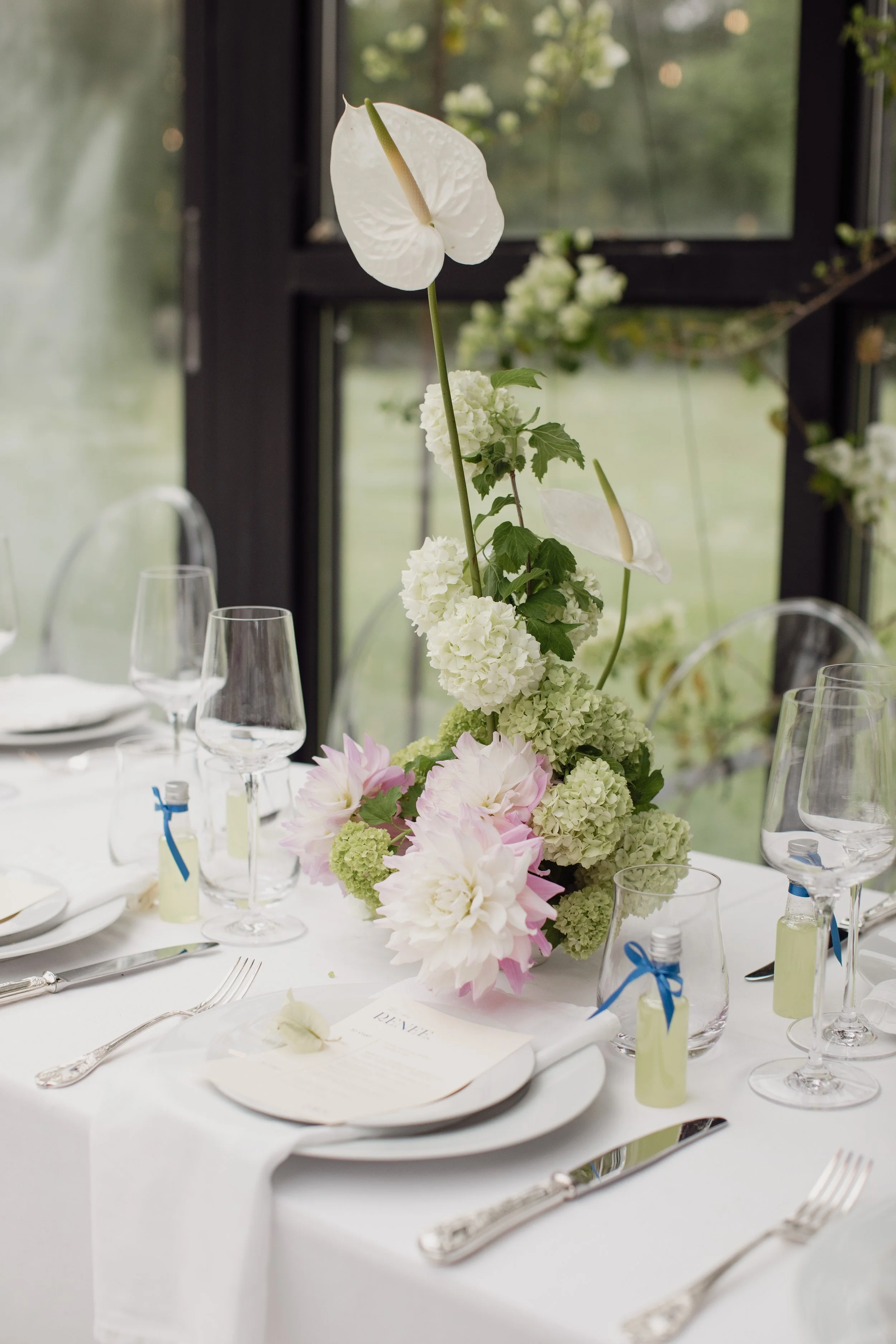 A formal dining table with a floral centerpiece containing white anthuriums, hydrangeas, and pink peonies. The table is set with white plates, silverware, wine glasses, and small bottles of water. There are menus or programs at each place setting and