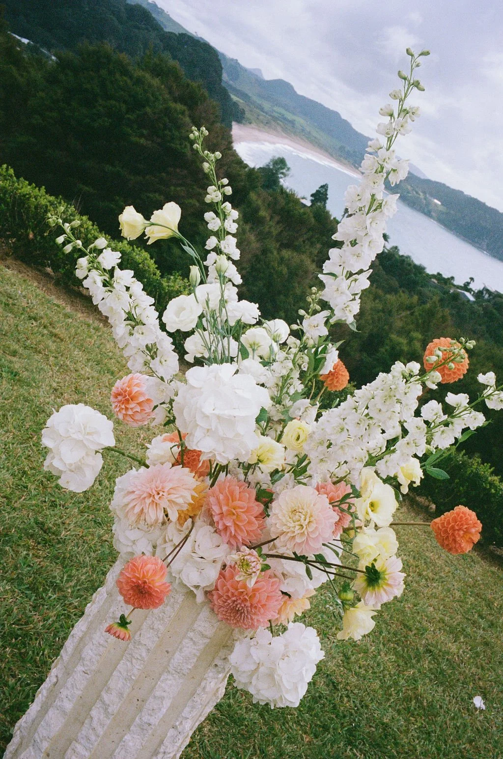A floral arrangement in a striped white vase featuring white, peach, and yellow flowers with a scenic landscape of green hills, water, and cloudy sky in the background.