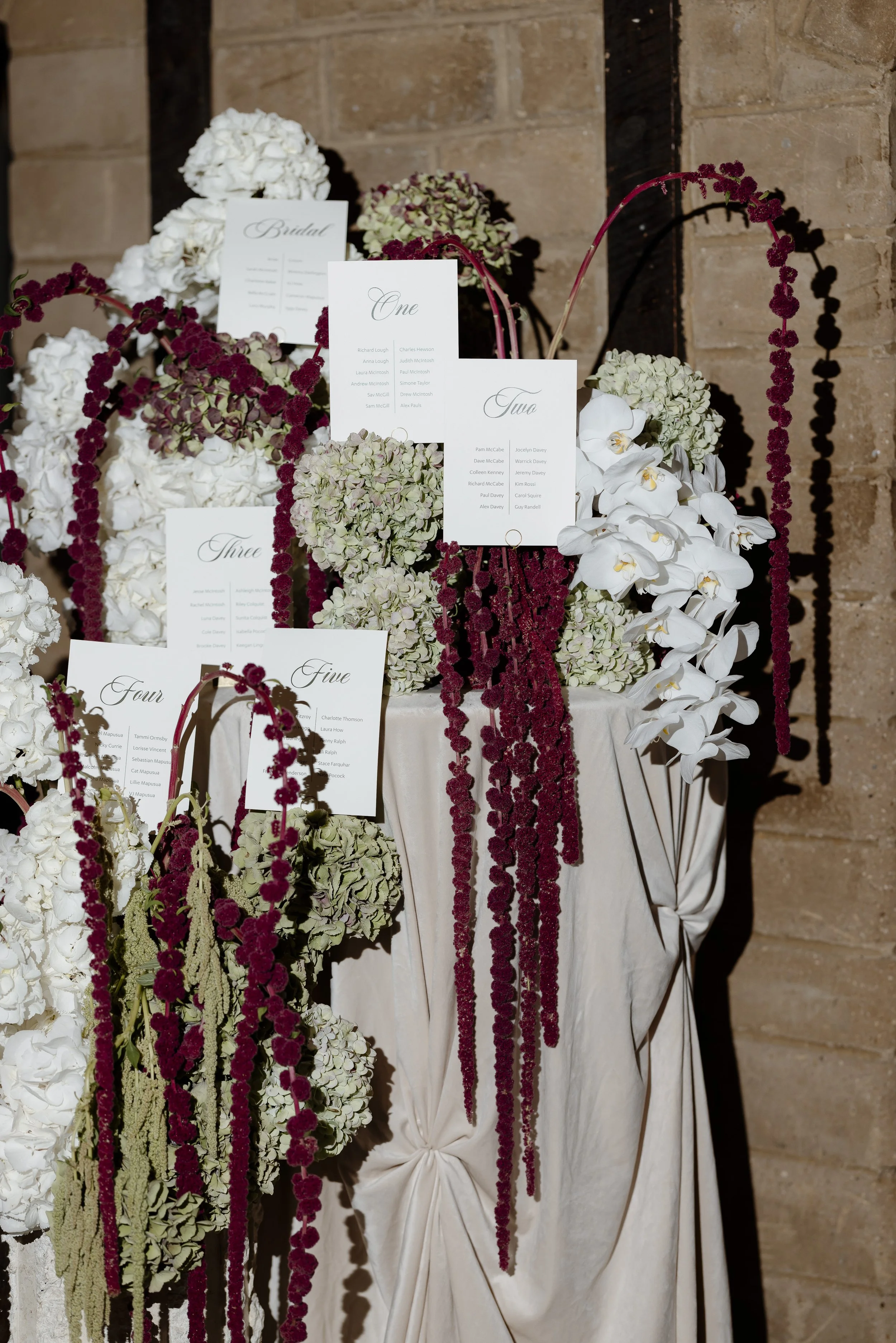 A floral table decoration with white and burgundy flowers and place cards for a wedding reception, set against a brick wall.