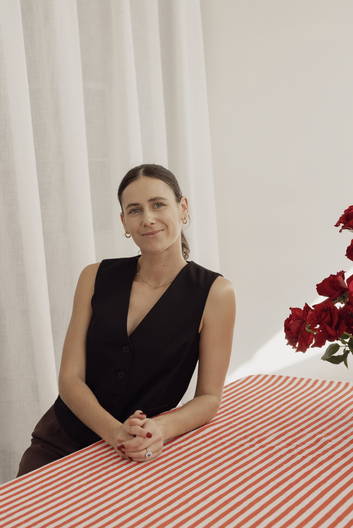 A woman with brown hair and blue eyes, wearing a black sleeveless vest, gold jewelry, and red nail polish, sitting at a table with a red and white striped tablecloth, a bouquet of red roses nearby, in a room with white curtains.