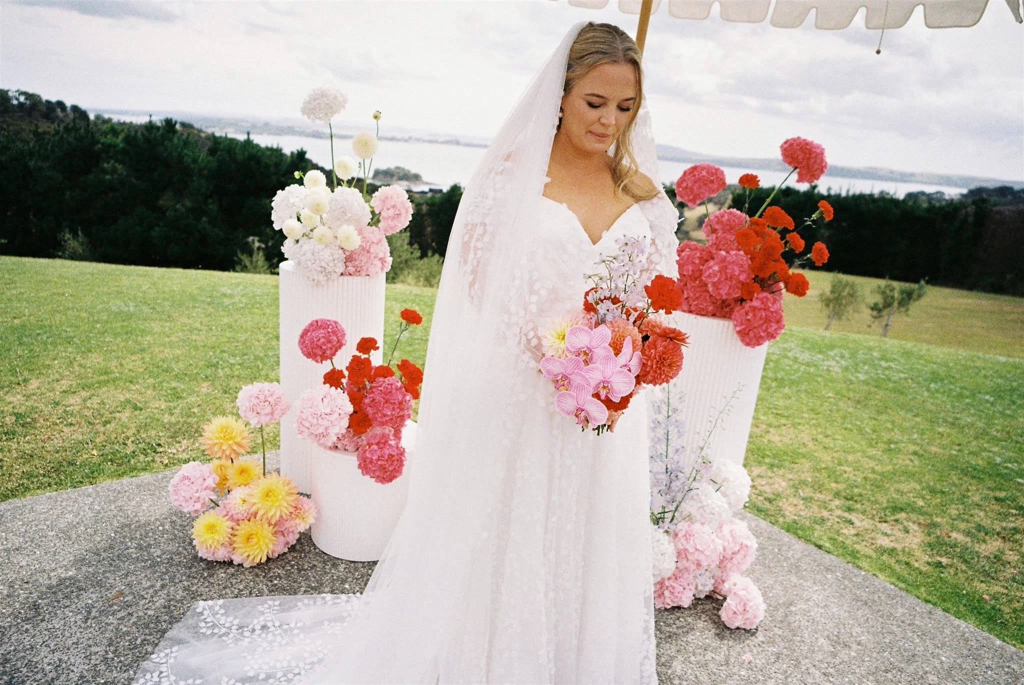 Bride holding her bright and colourful bouquet with her ceremony flowers behind her on plinths. Waiheke wedding.