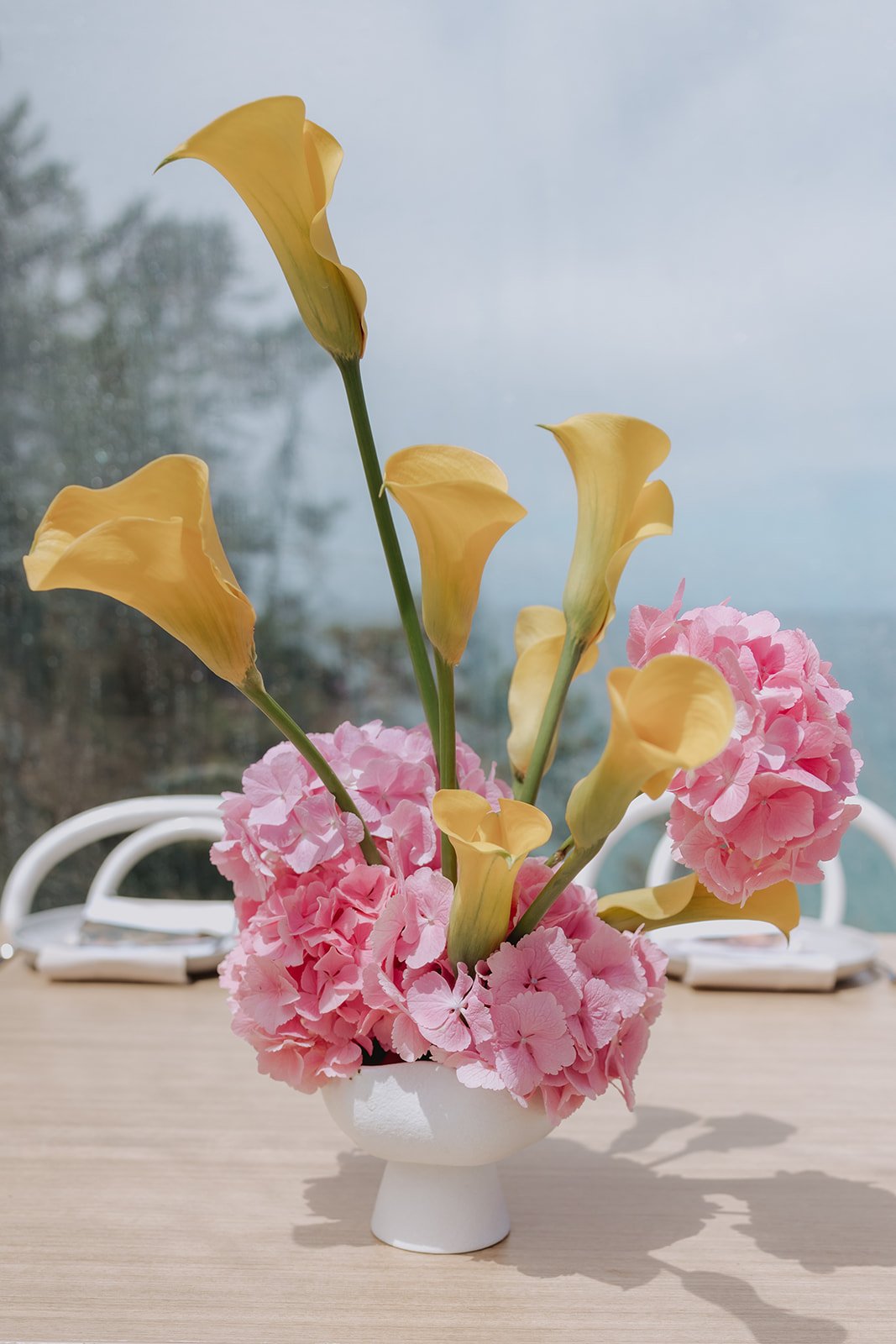 A white ceramic vase with pink hydrangea flowers and tall yellow calla lilies on a wooden table, with a foggy outdoor scenery in the background.