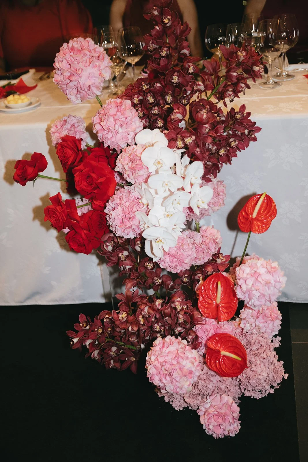 A floral centerpiece with pink hydrangeas, red roses, white orchids, and red anthuriums on a table at a formal event.