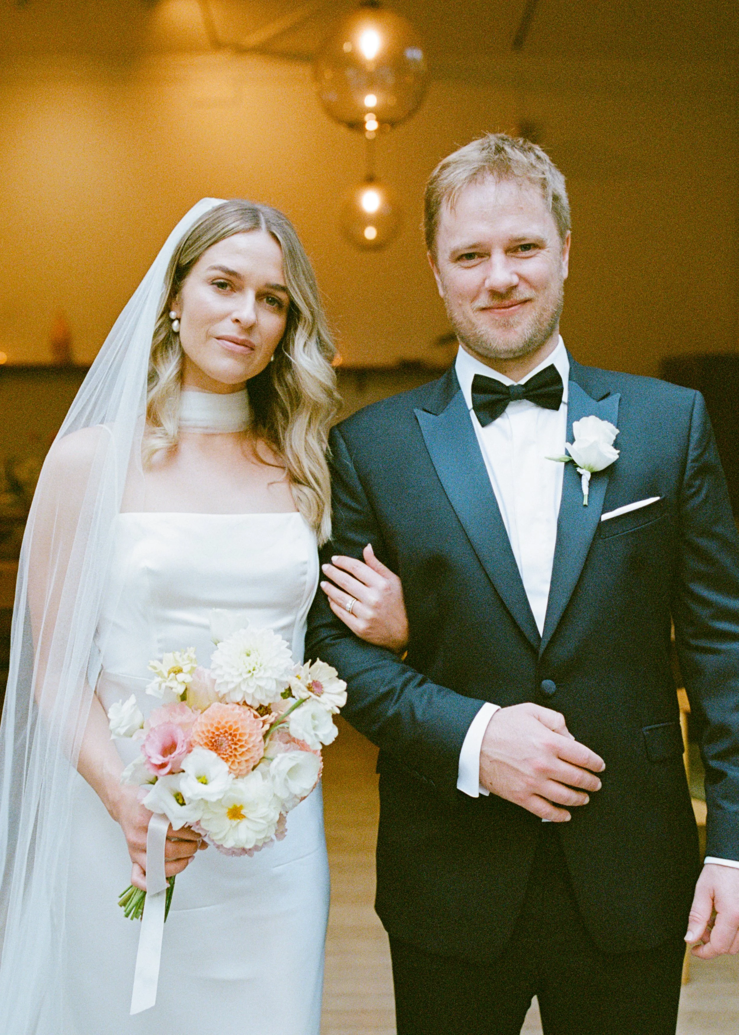 A bride and groom on their wedding day, standing close together indoors with warm lighting. The bride is holding a bouquet of pastel-colored flowers and wearing a white wedding dress with a veil. The groom is dressed in a black tuxedo with a black bo