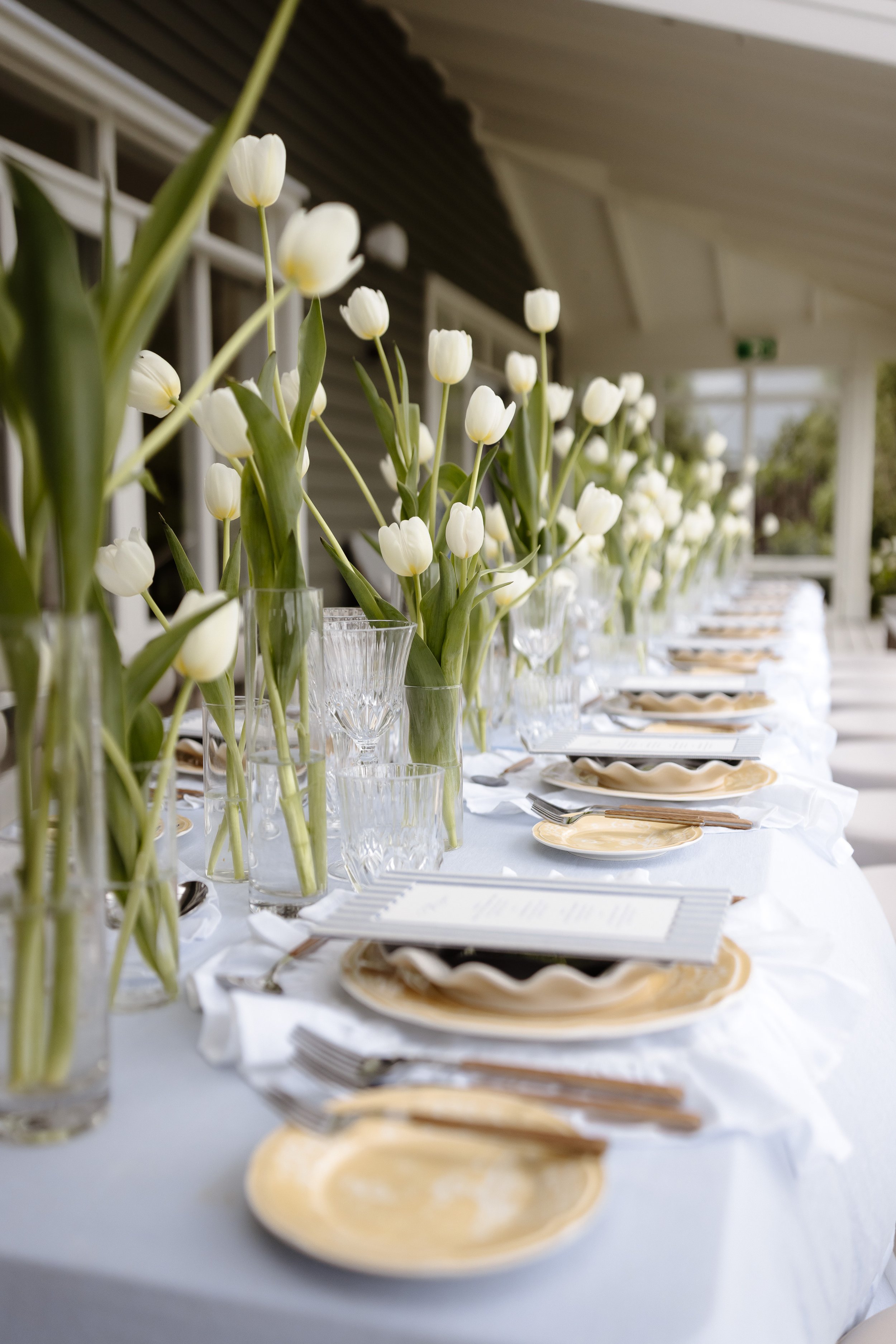 White tulips in glass vases arranged on a table with plates, forks, and glasses, set for a formal event or gathering on a covered porch or patio.