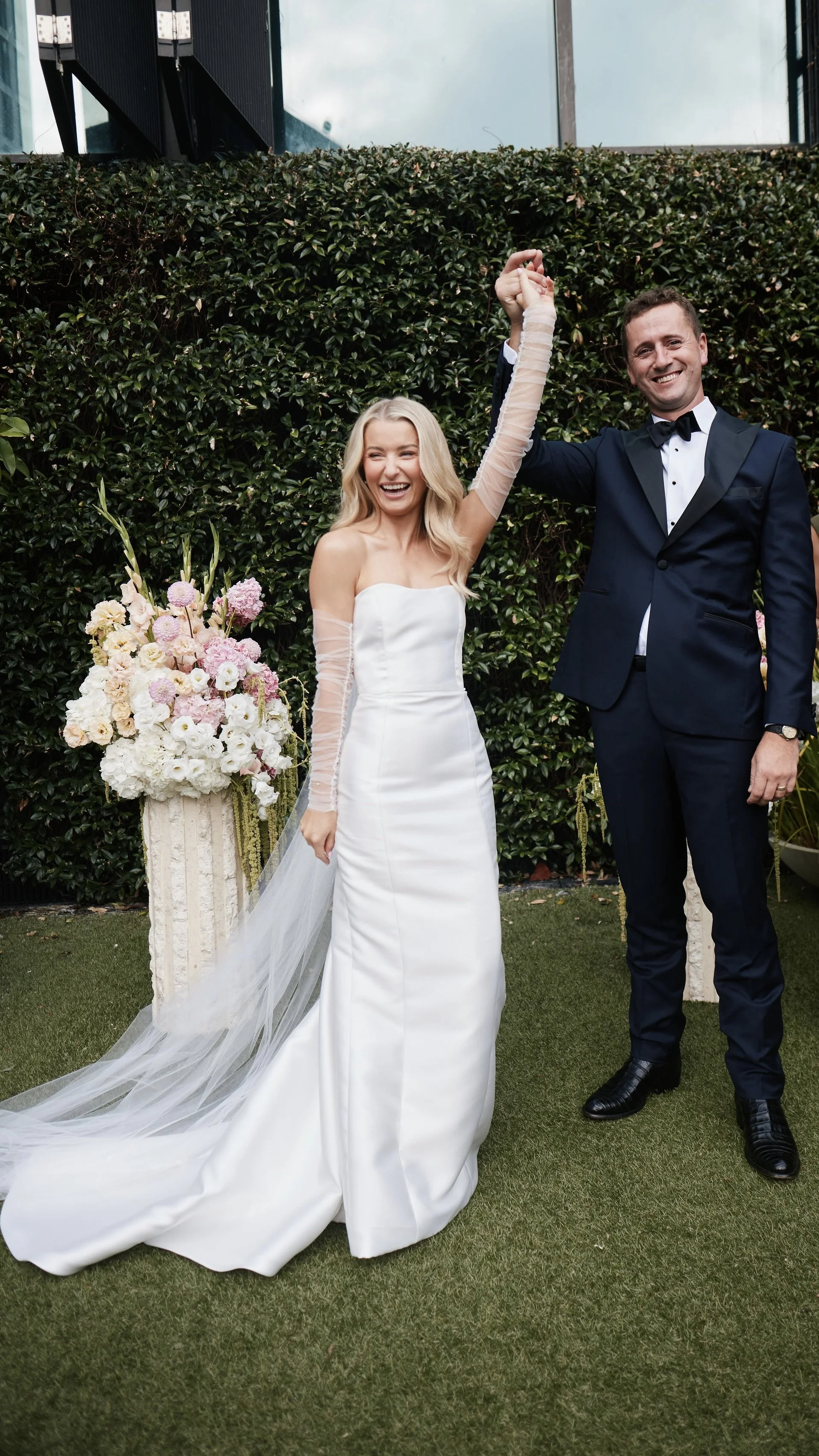 A bride and groom are celebrating outdoors on their wedding day, lifting their hands together. The bride is wearing a strapless white satin wedding gown with sheer long sleeves, and the groom is dressed in a navy tuxedo with a bow tie. There is a lar