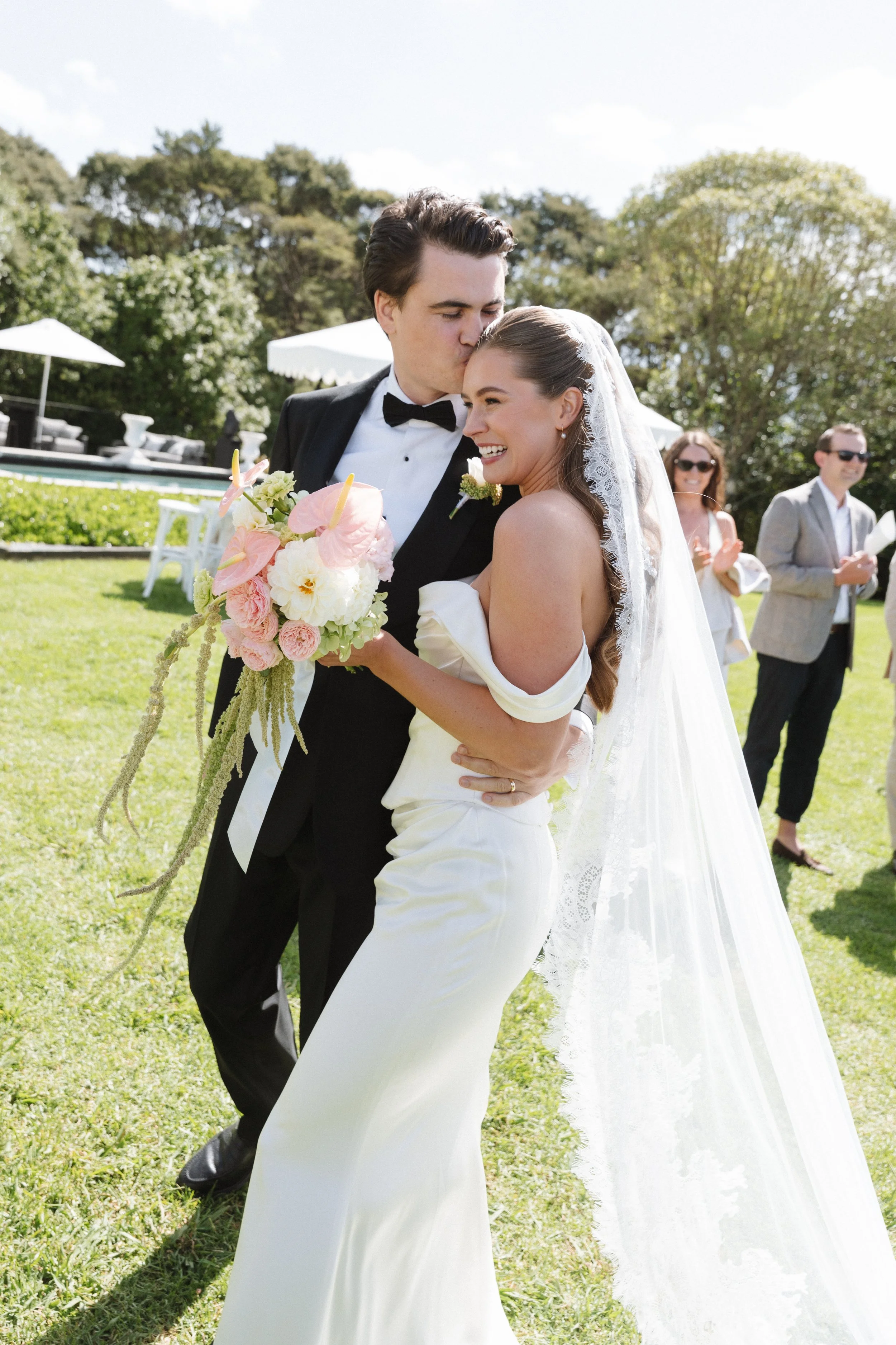 A newlywed couple, the groom in a tuxedo and the bride in a white wedding gown with a veil, sharing a joyful moment outdoors on a sunny day. The groom is kissing the bride on the forehead while she smiles warmly, holding a bouquet of pink and white f