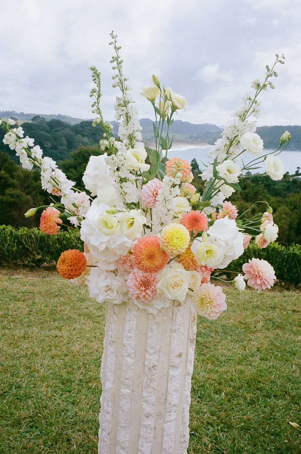 A tall white textured vase holding a mixed arrangement of white, peach, and yellow flowers outdoors, with a landscape featuring a body of water, hills, and cloudy sky in the background.