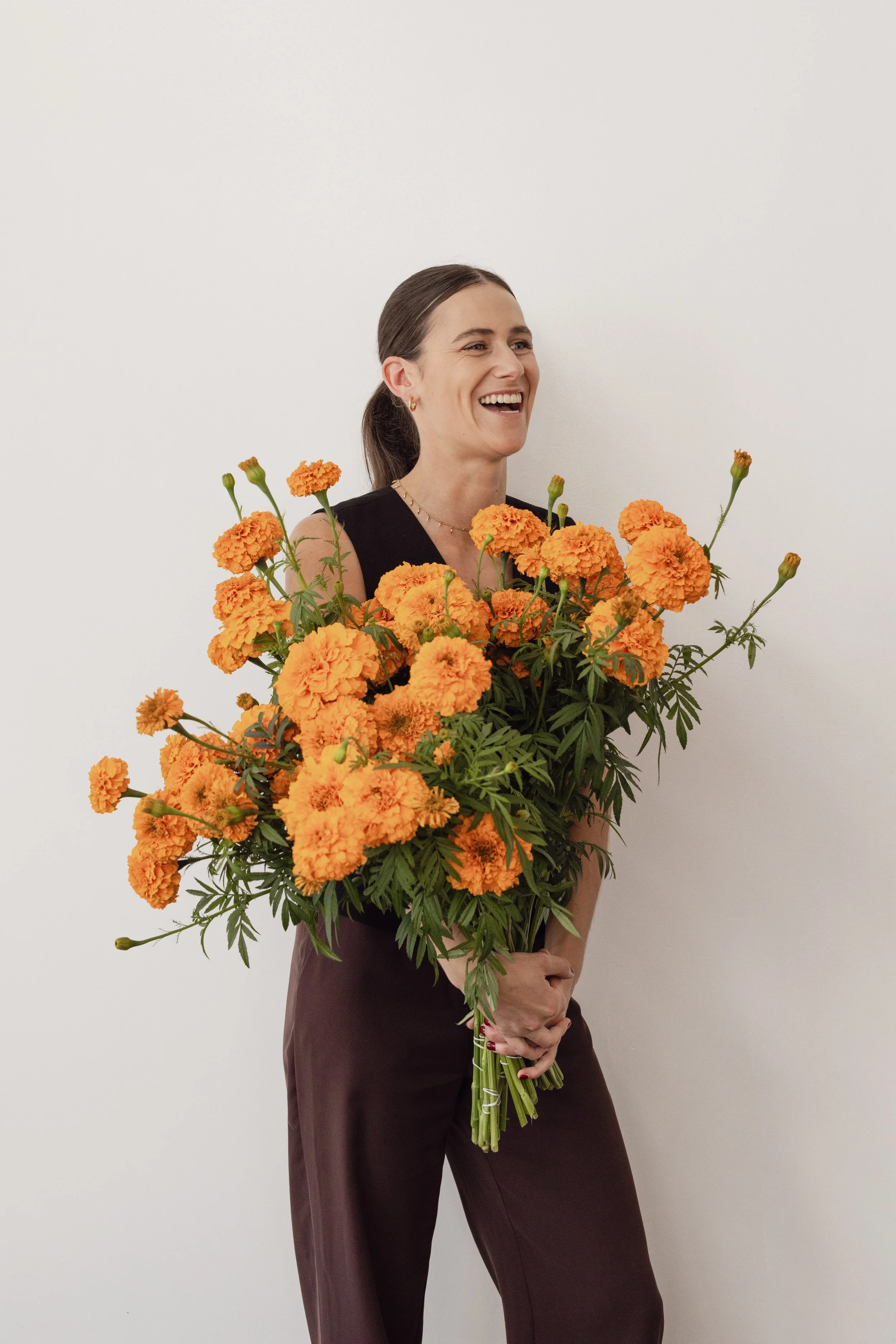 A woman holding a large bouquet of orange marigold flowers, standing against a plain white wall, smiling and looking to her right.