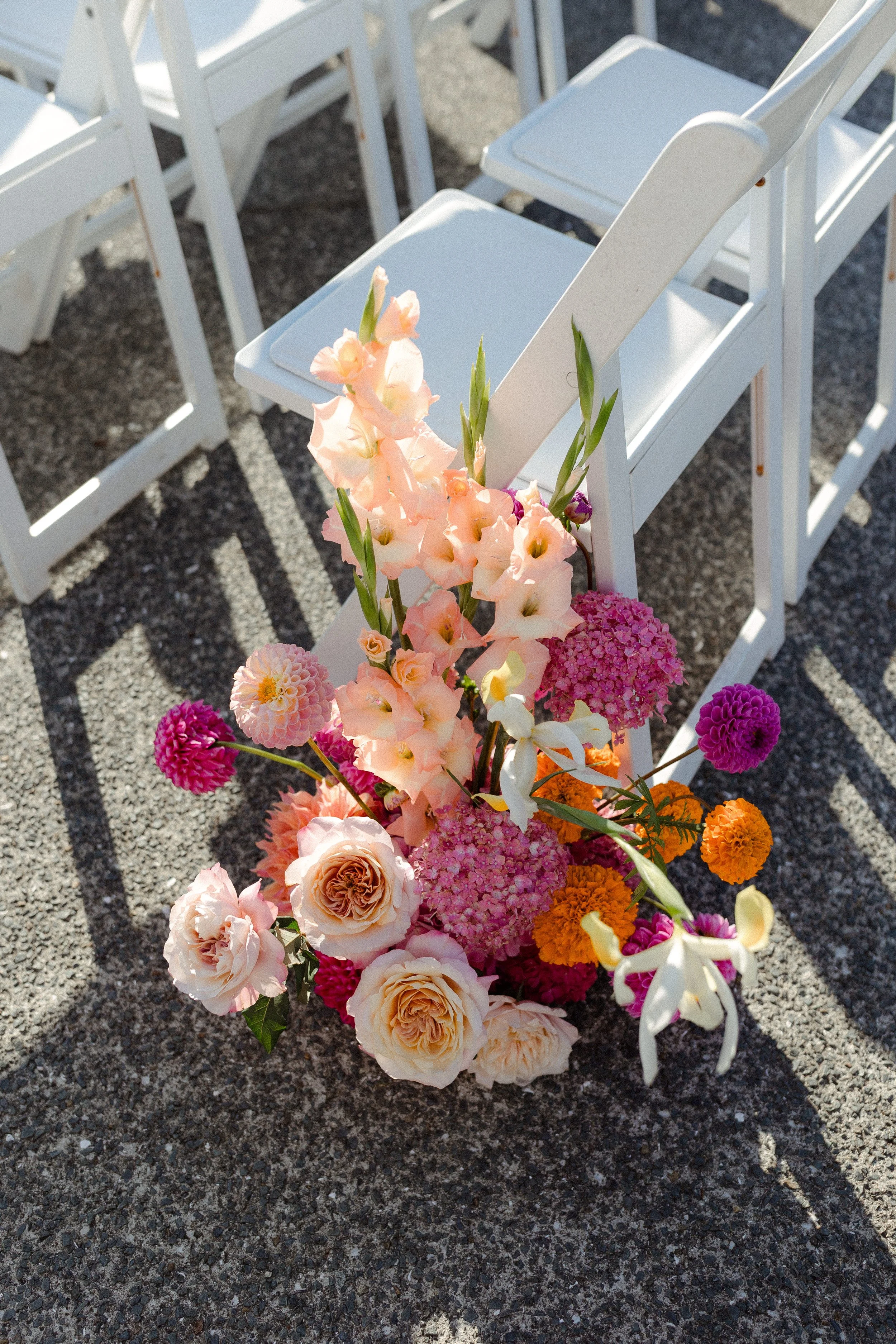 A group of white folding chairs with floral arrangements placed on some of them, on a gravel surface.