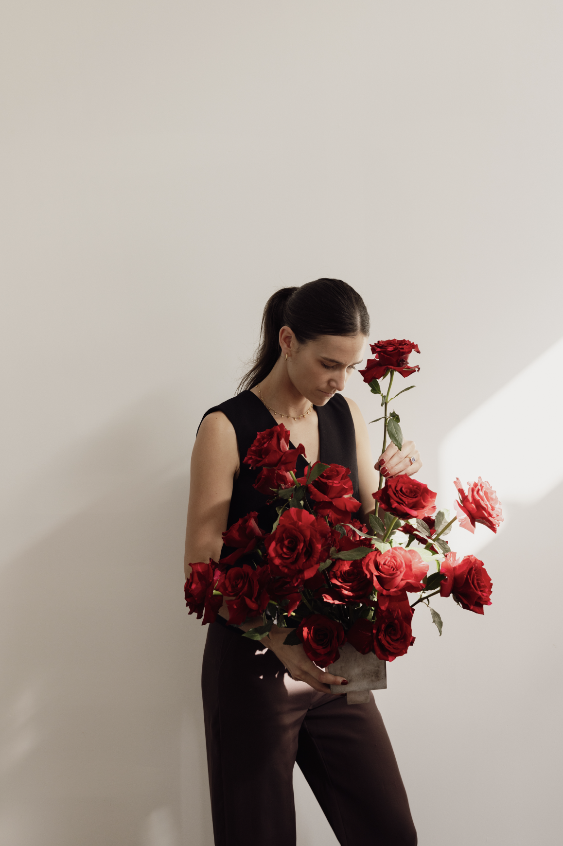 A woman with dark hair tied back, wearing a black sleeveless top and dark pants, holding a large bouquet of red roses against a plain white wall.