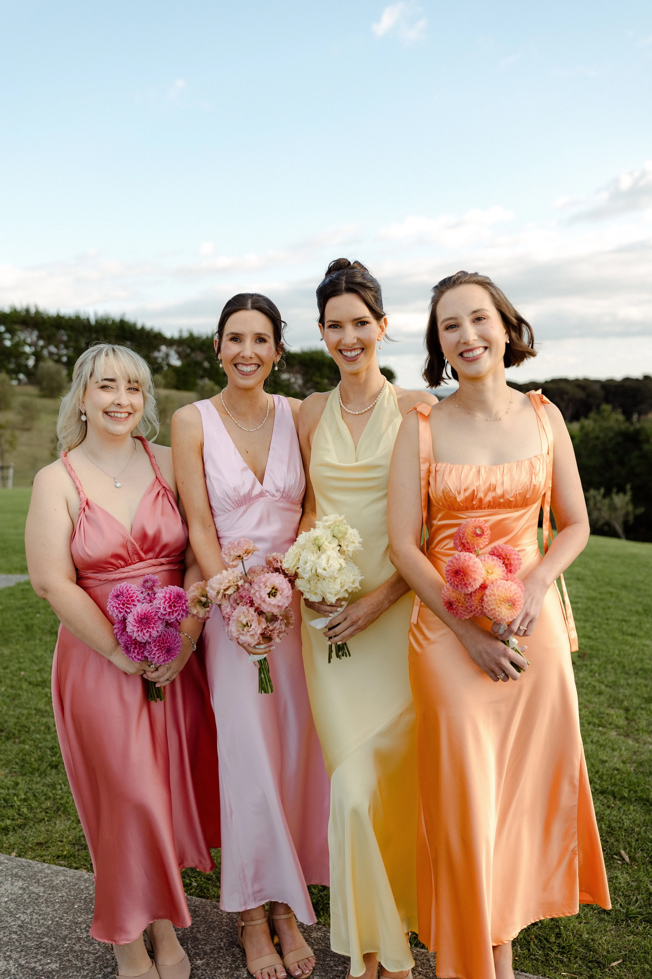 Four women in colorful dresses holding bouquets, standing outdoors on grass with trees and a partly cloudy sky in the background.