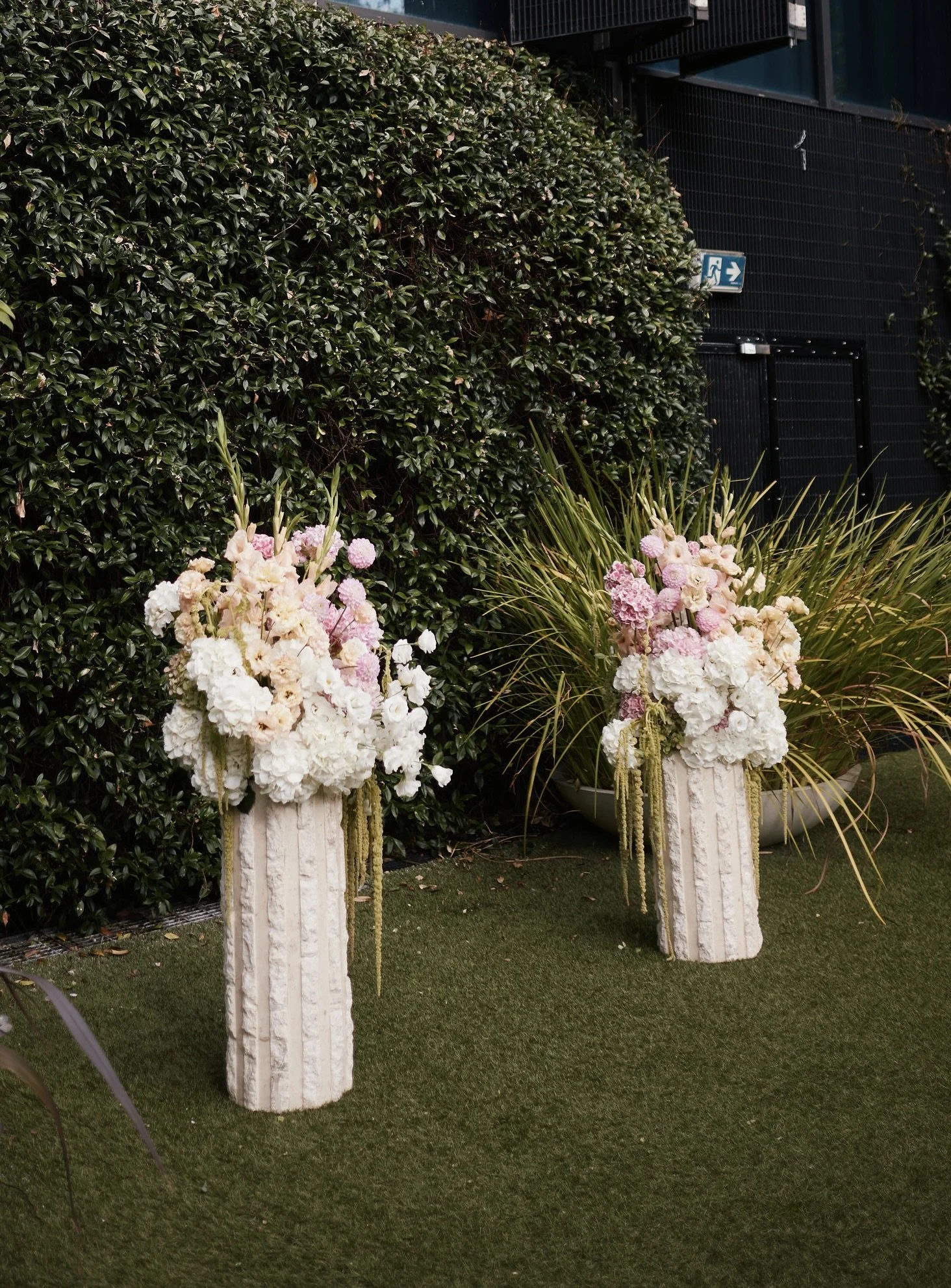 Two white and pink floral arrangements on tall white pedestal vases outdoors in front of a green hedge and dark building with an emergency exit sign.