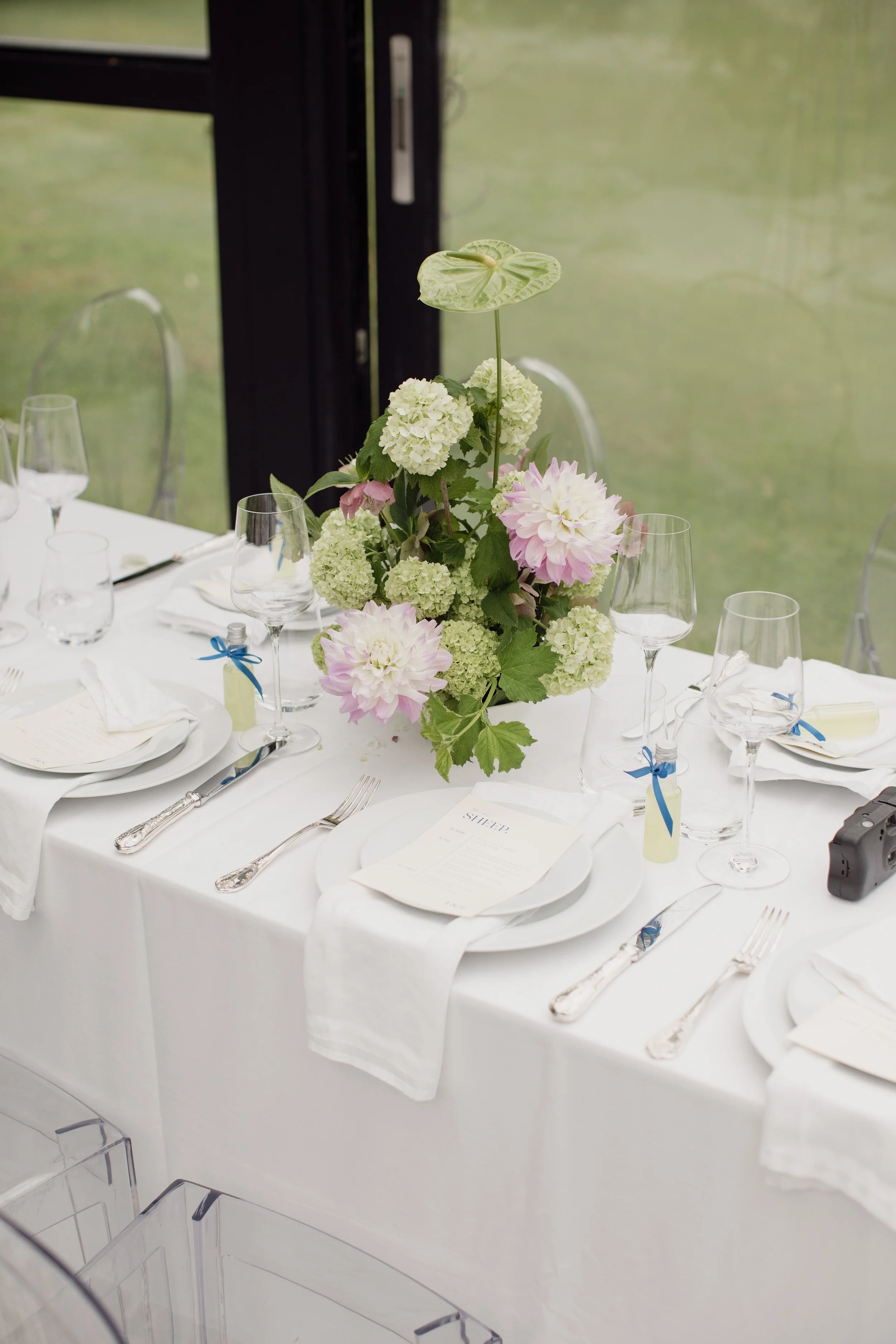 A table set for a formal event with a floral centerpiece featuring white and pink flowers. The table has white tablecloths, white plates, silverware, empty wine glasses, and small bottles with blue ribbons. There are also menus on the plates and a wi