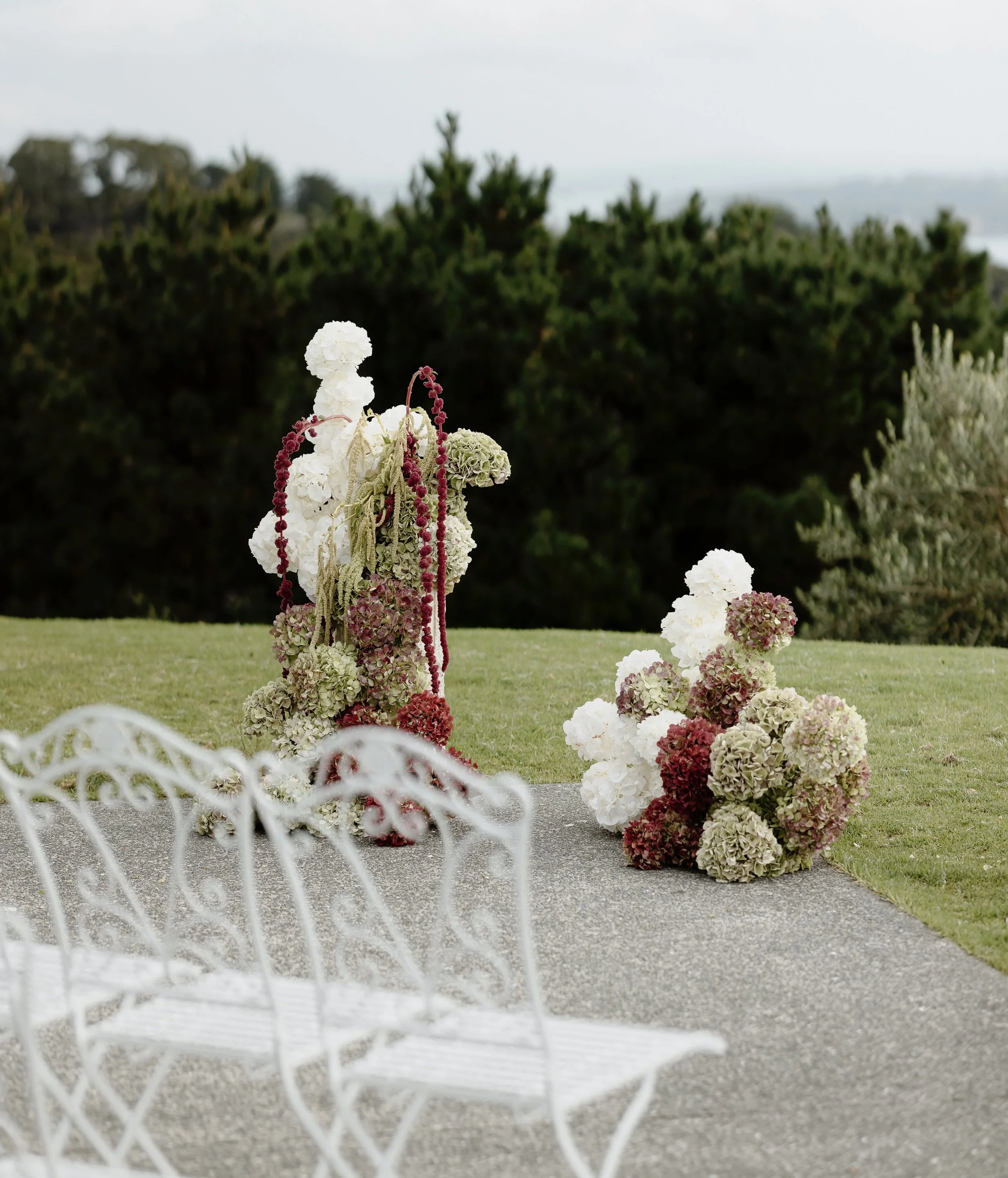 Decorative floral arrangements consisting of white, pink, and green hydrangeas and trailing red and beige plants on a grassy surface, with a blurred green hedge and cloudy sky in the background, seen through a white wrought iron fence.
