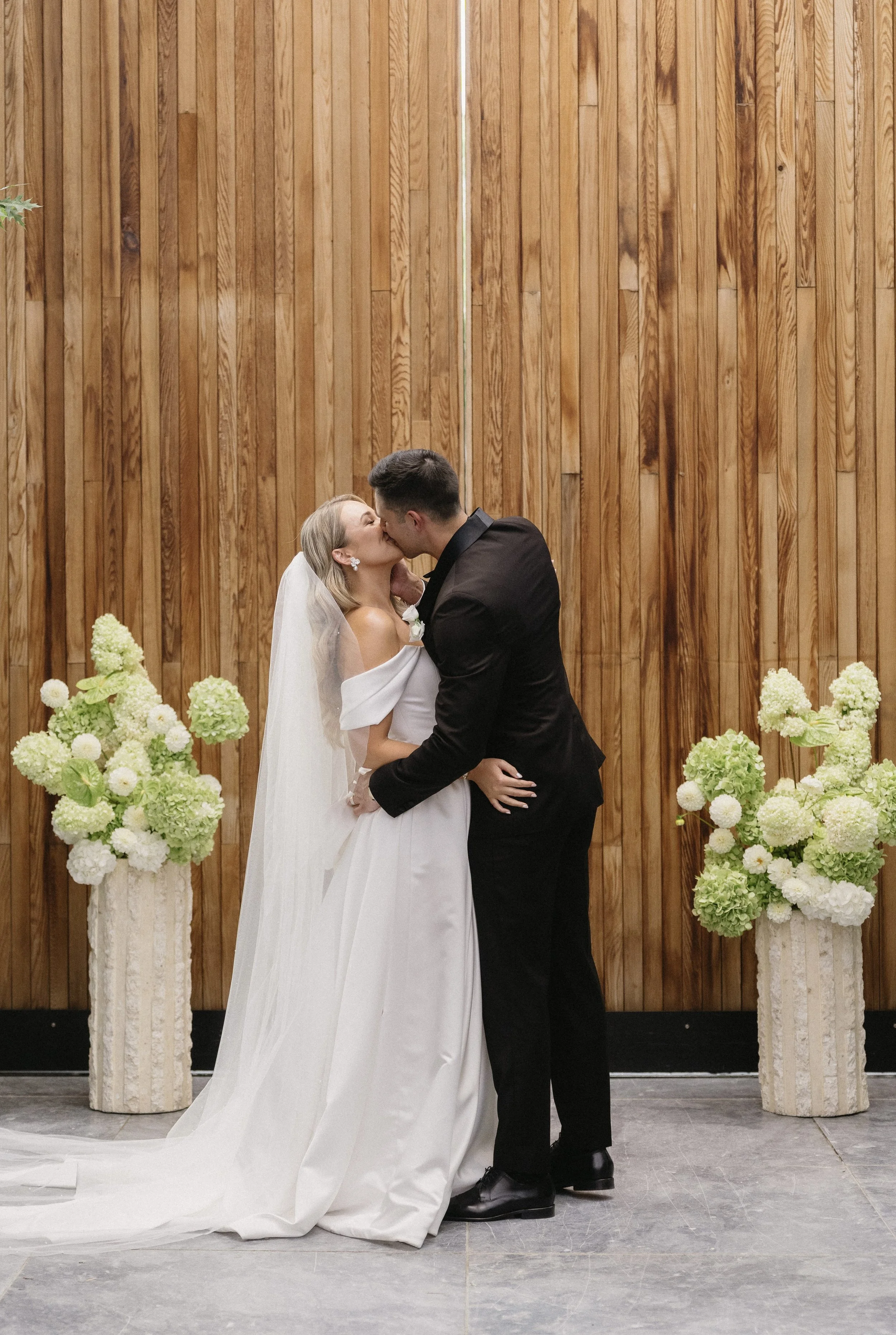 A bride and groom kissing during their wedding ceremony, standing in front of a wooden backdrop and floral arrangements.