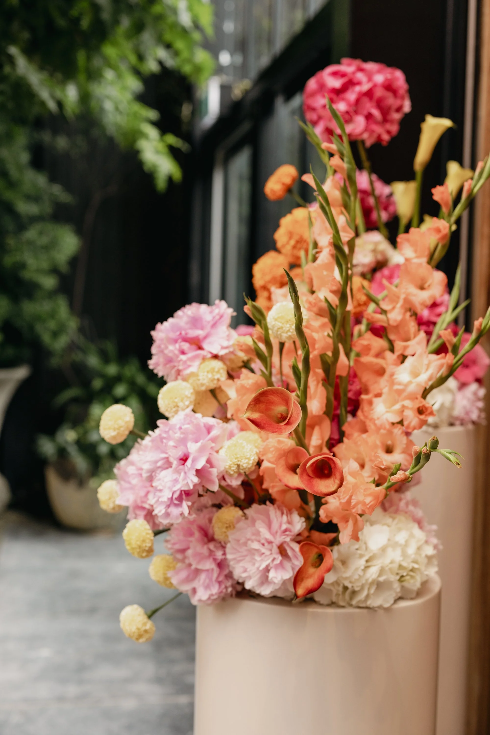 A white vase filled with a large, colorful flower arrangement including pink, peach, orange, and white flowers, placed on a gray surface near a wooden wall and a window with a glass pane.