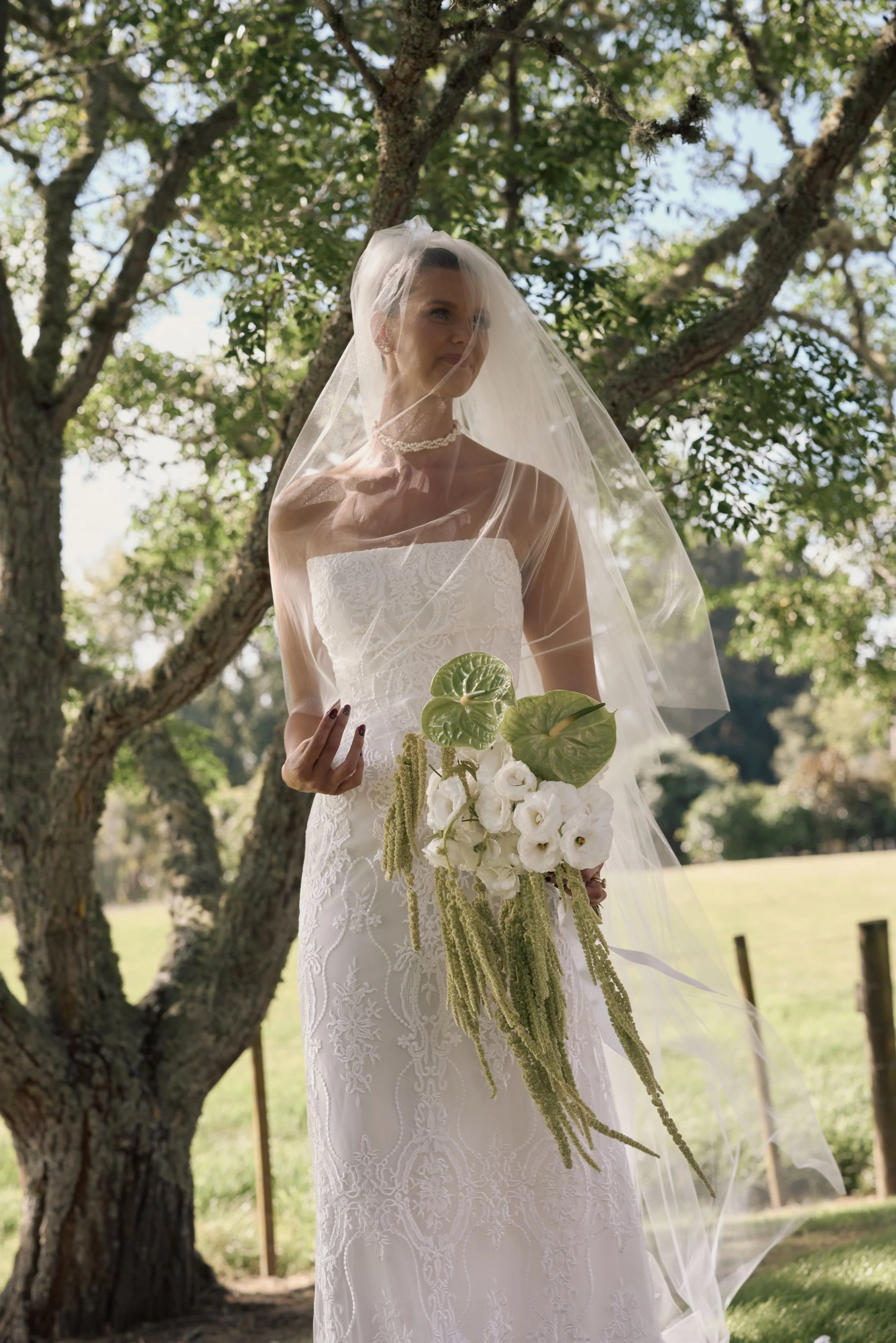 A bride in a white wedding dress with lace details, wearing a pearl necklace and a veil, stands outdoors in a park with trees and sunlight.