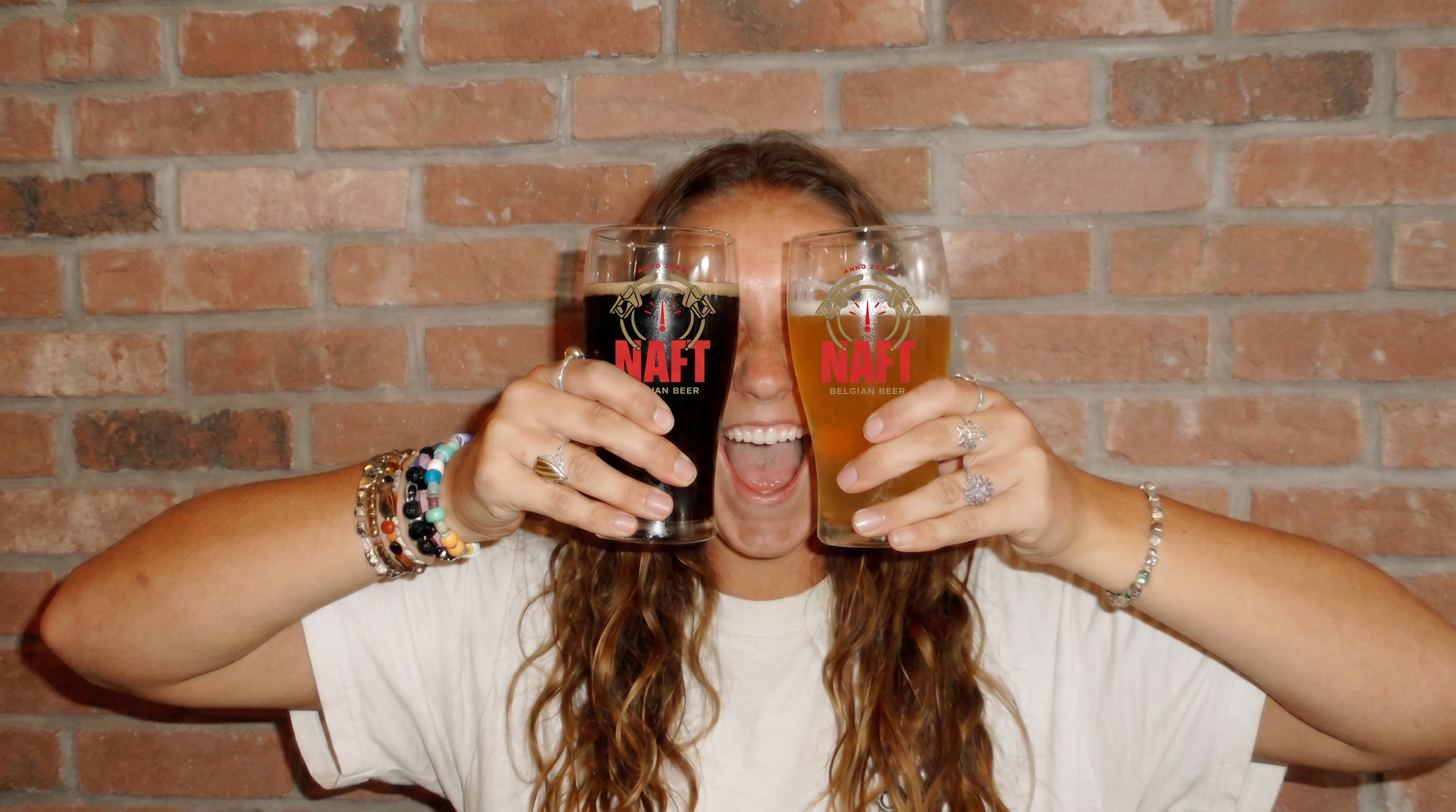 Vrolijke vrouw met lang, krullend haar die twee glazen bier vasthoudt, een met donker bier en een met licht bier, voor een bakstenen muur. Mensen drinken een naft bier. Ipa naft.