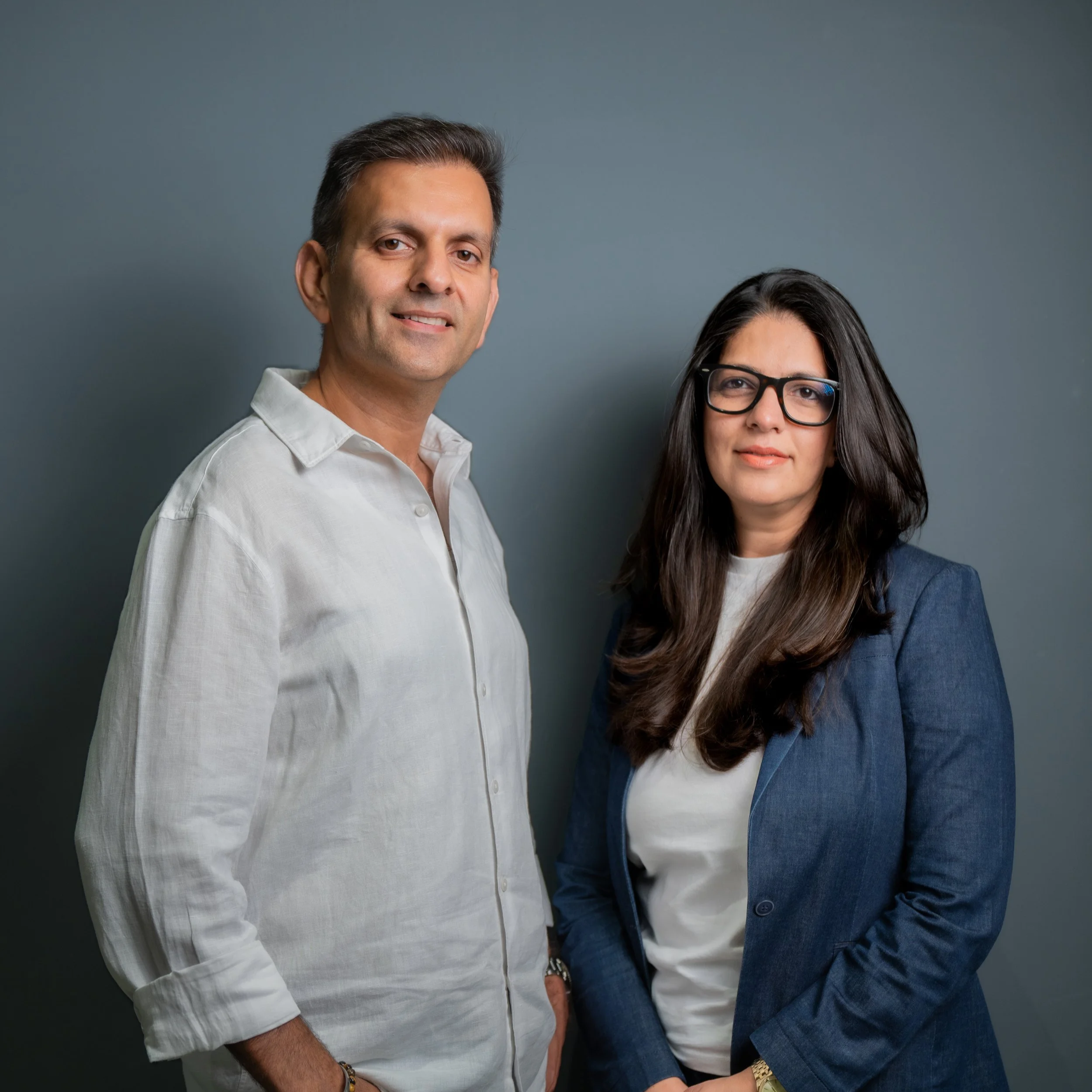 A man and a woman standing against a plain dark background. The man has short dark hair, is smiling slightly, and is wearing a light-colored shirt and a bracelet. The woman has long dark hair, glasses, and is wearing a denim jacket over a white shirt.