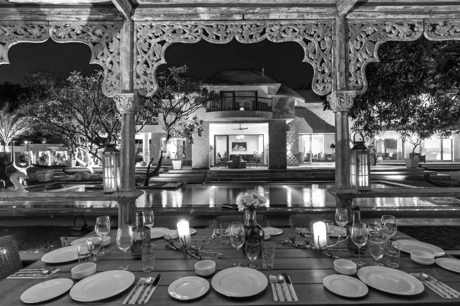 Black and white image of a beautifully decorated outdoor dining area with a table set with plates, glasses, and silverware. The scene overlooks a pool with trees, lanterns, and a house in the background, illuminated at night.