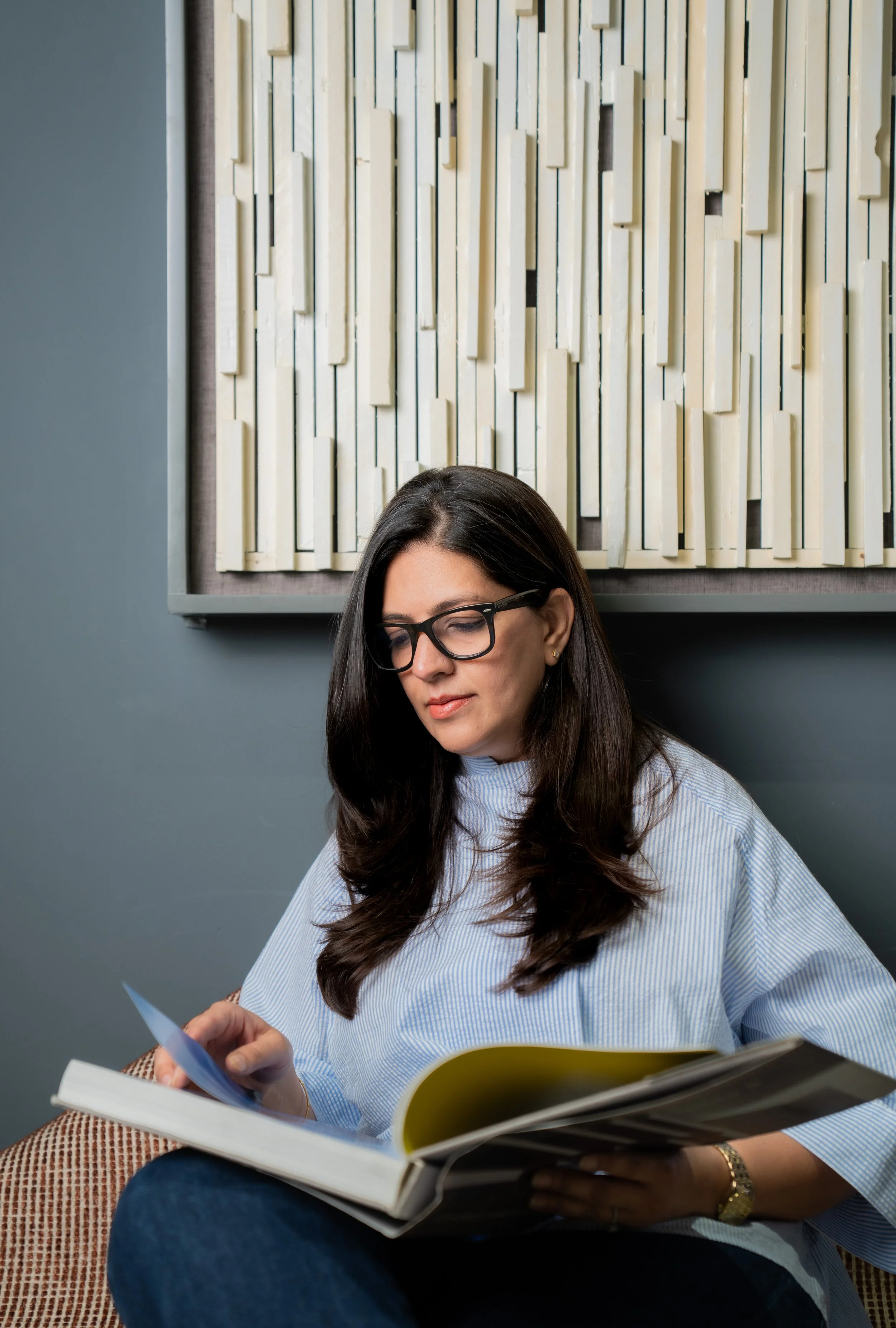 A woman with long dark hair, glasses, and a blue striped shirt sitting on a chair, reading a book and holding papers, in a room with a decorative beige and cream wall art in the background.