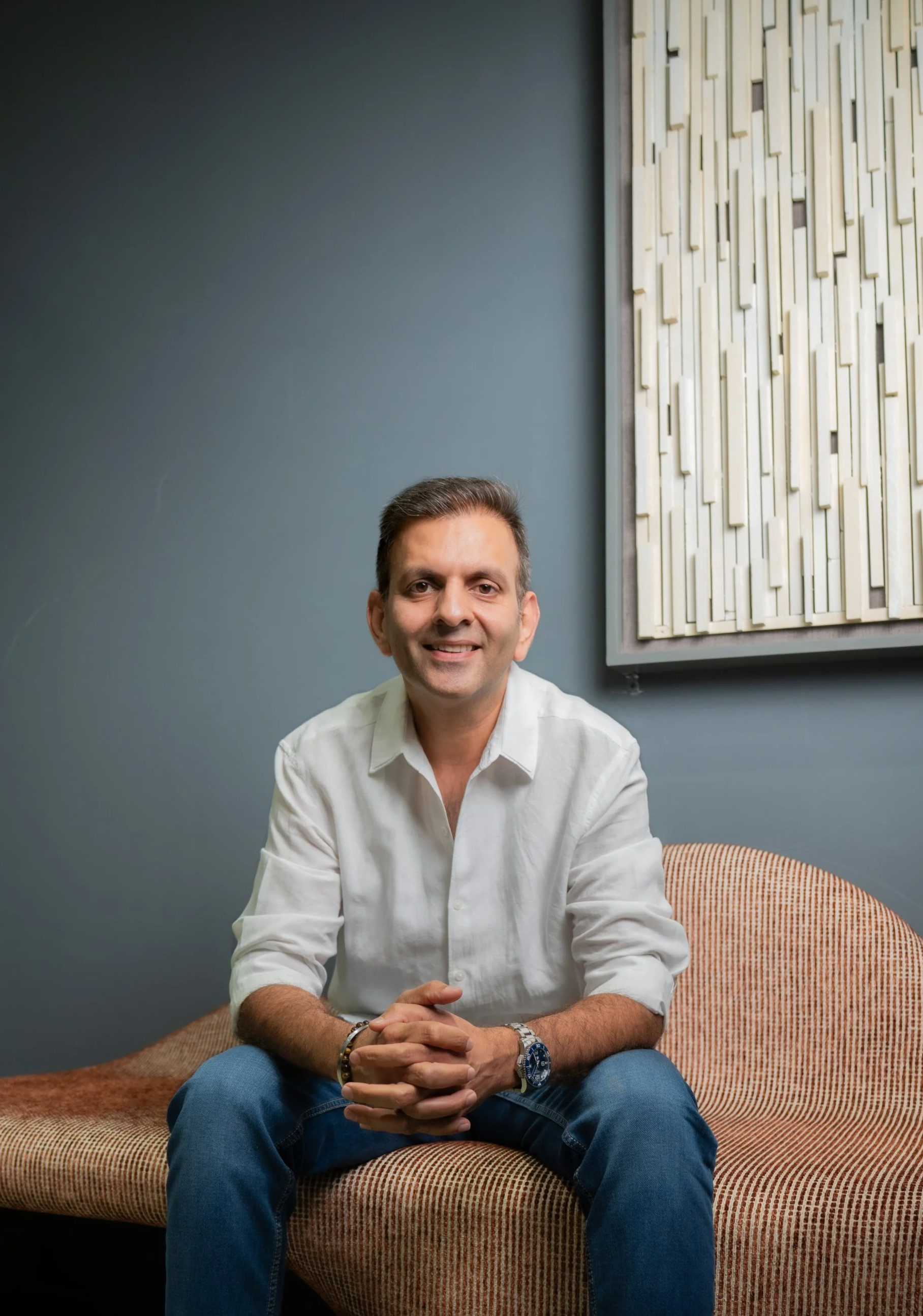 A man with short dark hair, wearing a white button-up shirt and blue jeans, sitting on a patterned orange and beige sofa in front of a blue wall with a wooden acoustic panel, smiling at the camera.