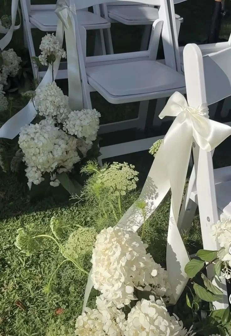 Chaises blanches décorées avec des rubans en satin crème et entourées de fleurs blanches, probablement pour un mariage.