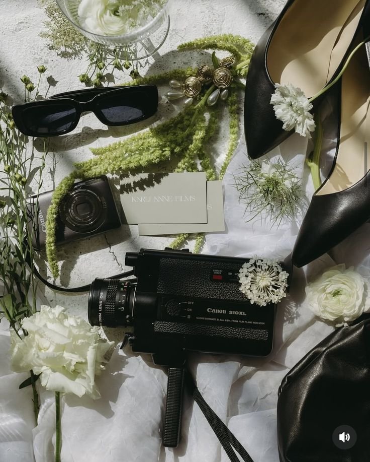 Objets divers sur une surface blanche, comprenant des lunettes de soleil noires, une caméra compacte, une clé USB, un appareil photo, une boîte de cartes de visite, des chaussures noirs à talons avec une fleur blanche, des fleurs blanches et vertes, et un verre d'eau.