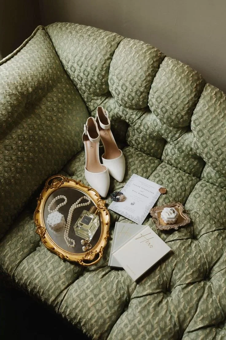 Une paire de chaussures blanches de mariage, un livre d'or, des bijoux, une bouteille de parfum, des invitations et une boîte à bijoux sur un canapé vert en velours.