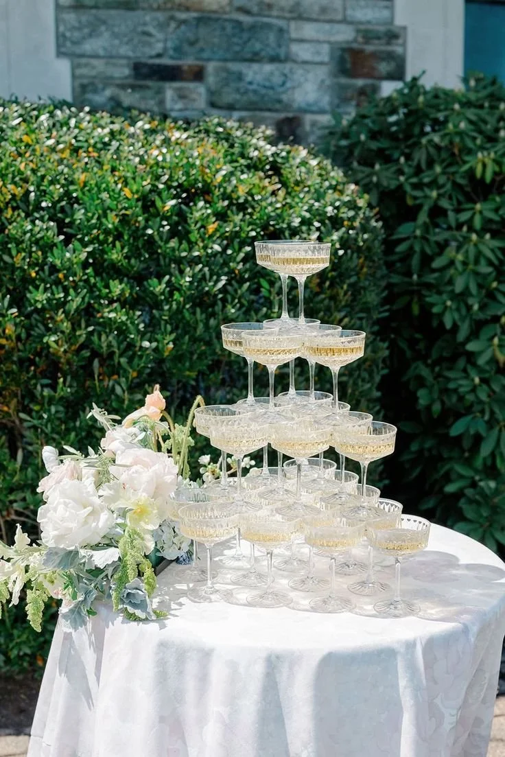 Un tas de coupes à champagne empilées en pyramide sur une table blanche, décoration florale à côté, en plein air avec des buissons verts en arrière-plan.