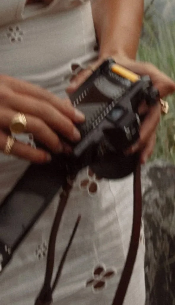 Une personne qui tient une arme à feu, probablement un pistolet, avec les mains visibles, portant plusieurs anneaux en or. La personne porte une robe ou une chemise légère avec des motifs de trous. L'image est floue.
