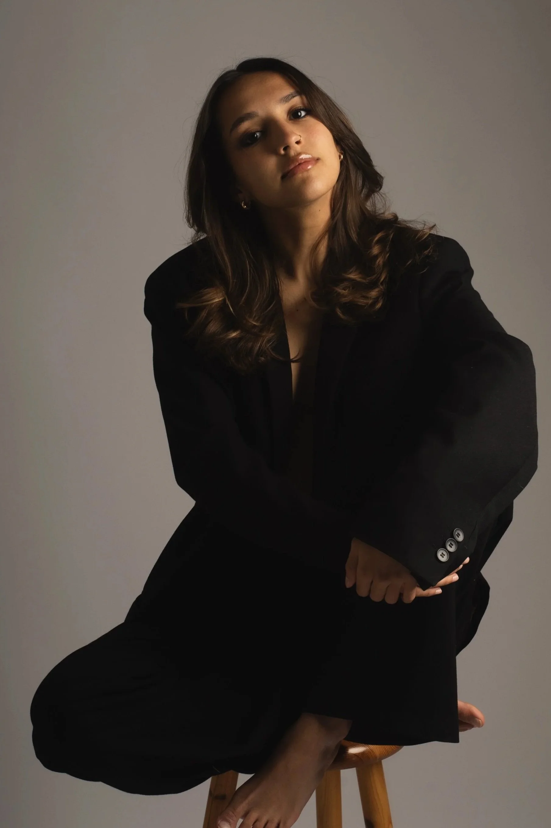 Une femme avec des cheveux bruns et ondulés, portant un blazer noir, posant devant un fond neutre, assise sur un tabouret en bois.