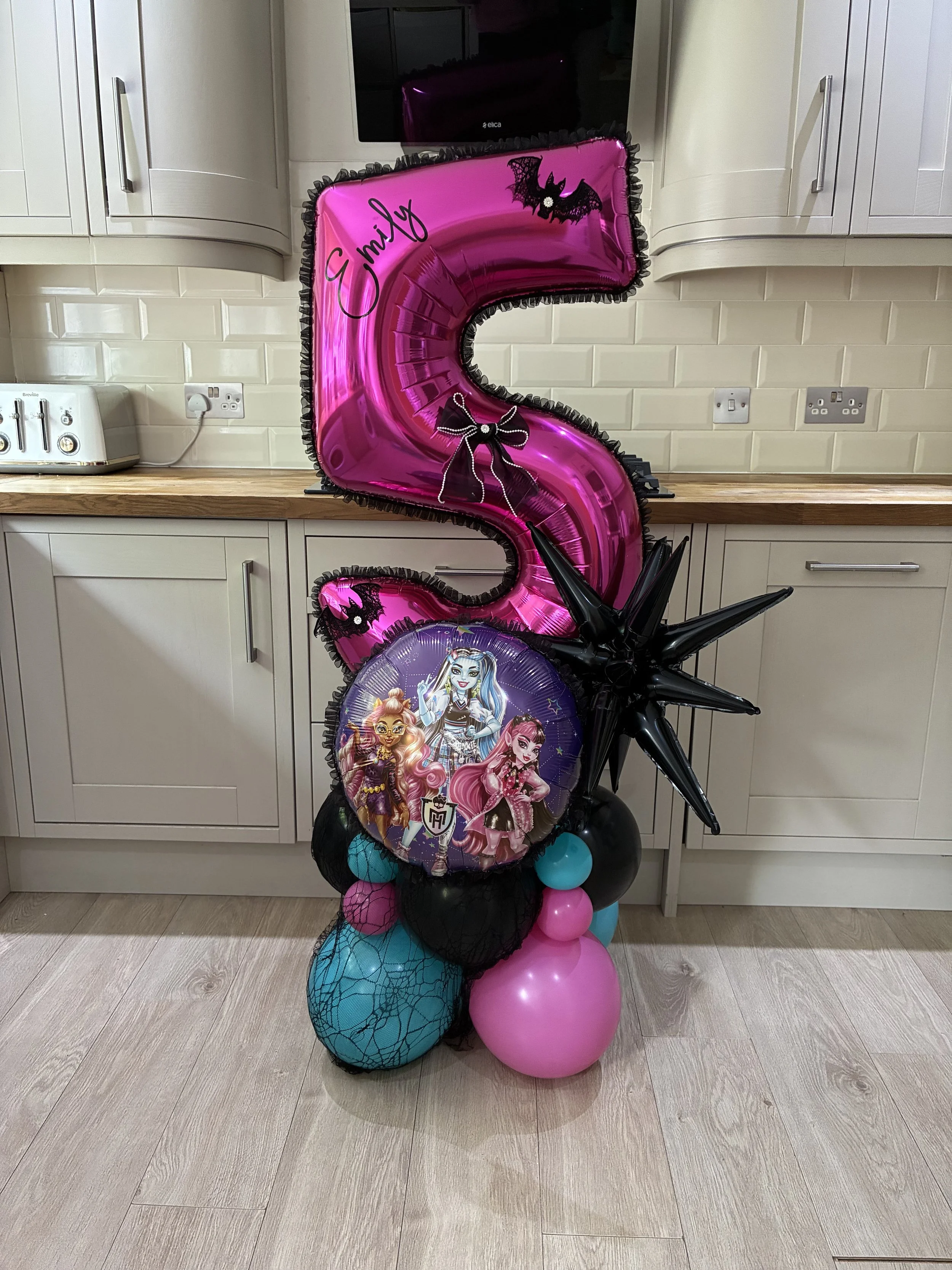 Colorful birthday balloon decoration featuring a large pink number 5 balloon with a black bat design, a round balloon with animated girl characters, black star-shaped balloons, and various pink, black, and teal balloons arranged on a kitchen floor.