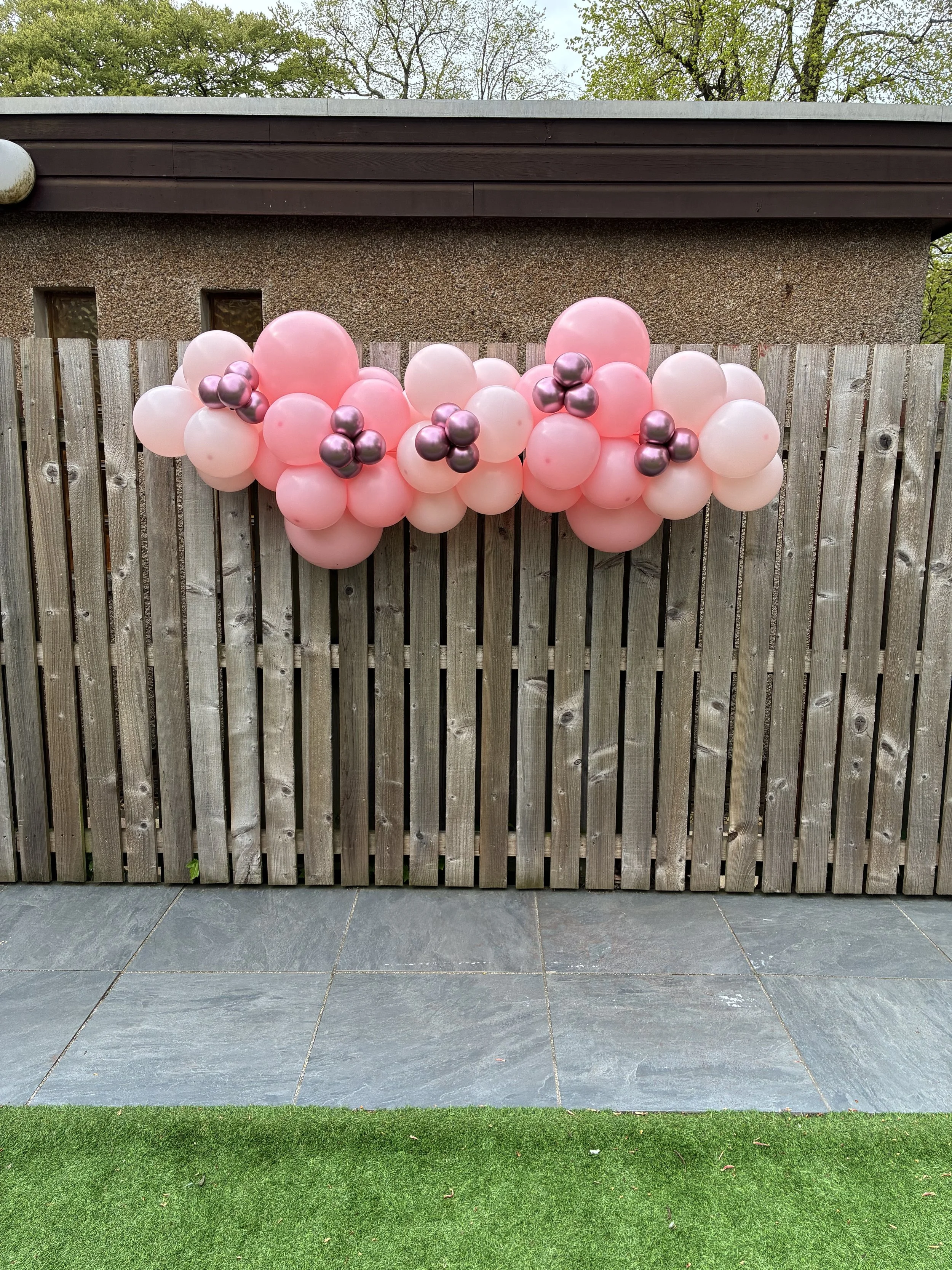 Pink and purple balloons arranged in clusters on a wooden fence outdoors, with a building, trees, and a paved ground in the background.