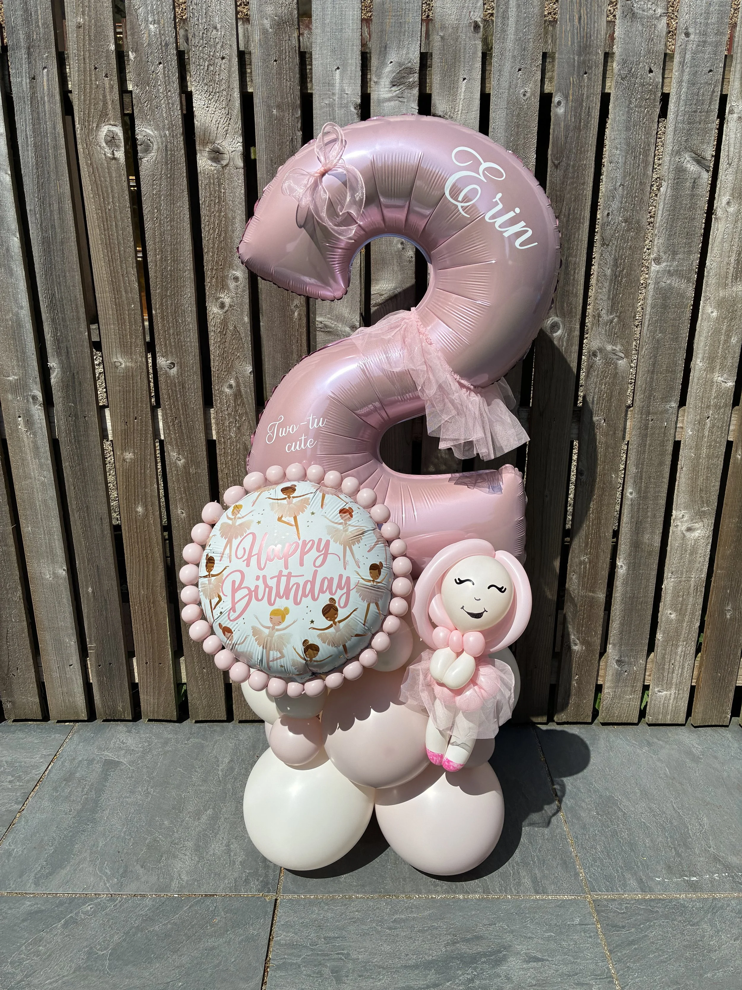 Pink ballet-themed balloons with the number 2, a "Happy Birthday" message balloon with ballerinas, and a smiling ballerina plush toy, set against a wooden fence background.