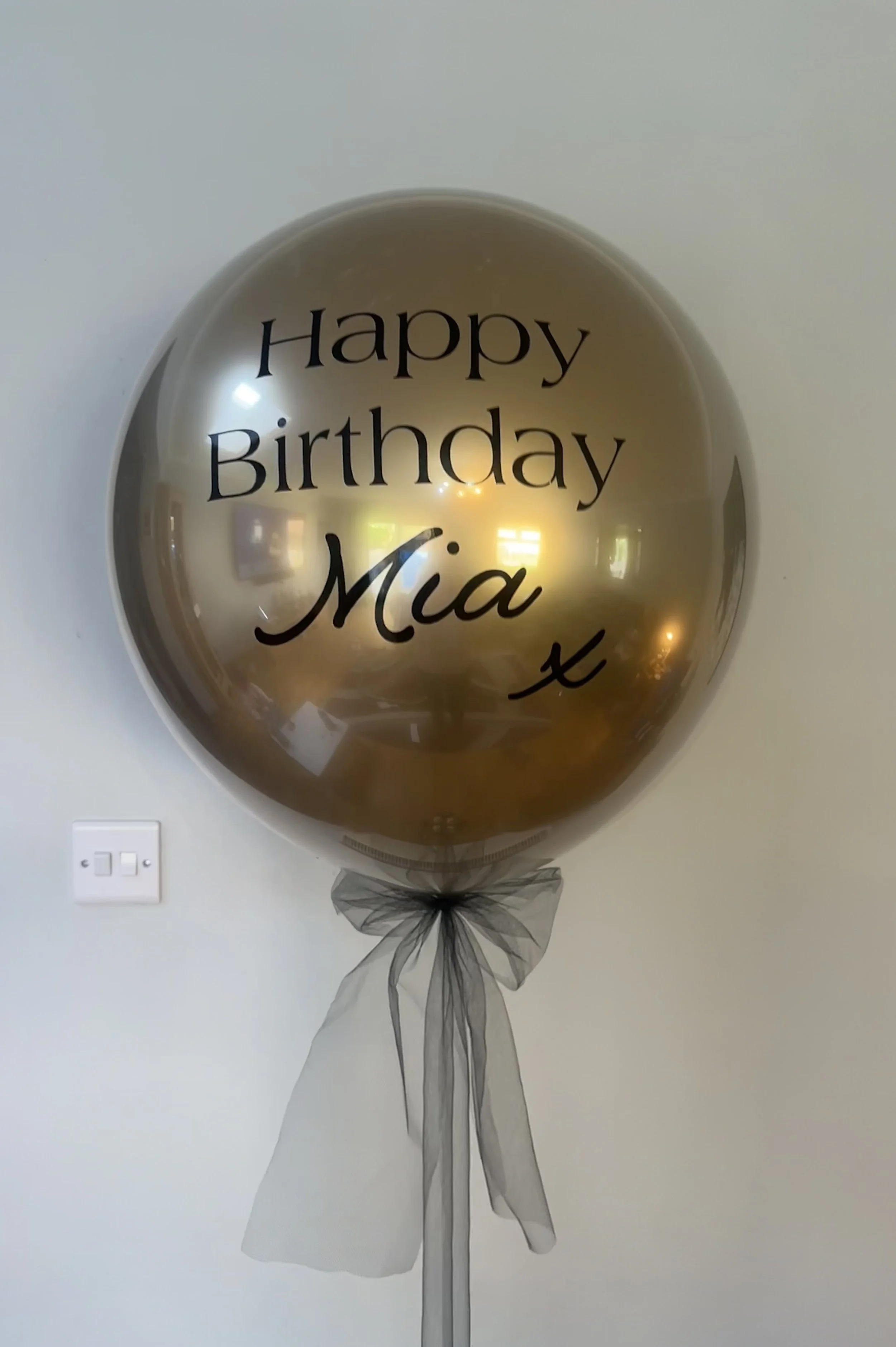 Gold birthday balloon with black text that reads, 'Happy Birthday Mia,' tied with black tulle on a white wall.