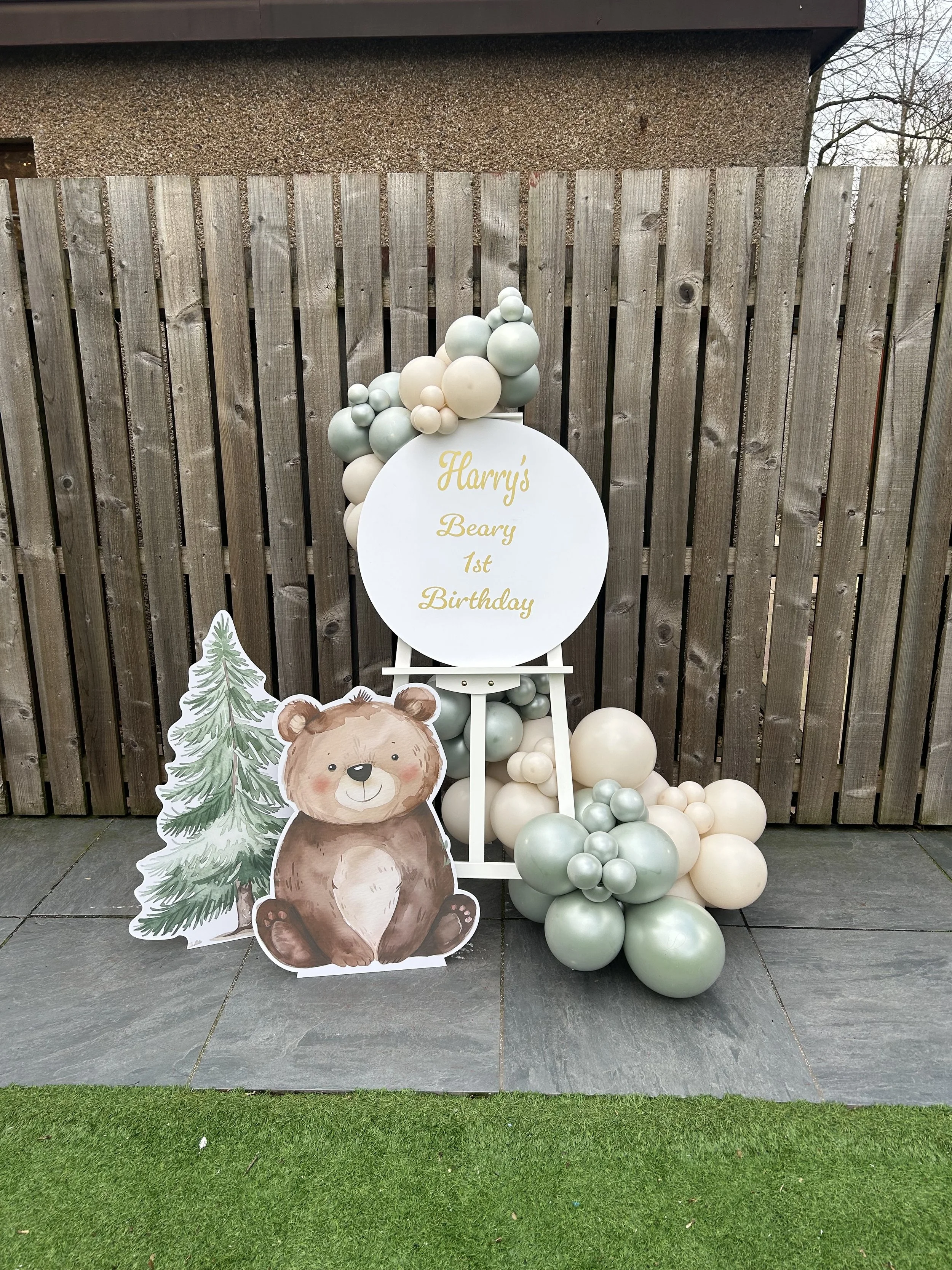 Decorative display for a child's birthday with a plush bear, a pine tree cutout, a white circular sign with gold text that reads "Harry's Beary 1st Birthday," and a balloon arrangement in shades of white, beige, and light green.